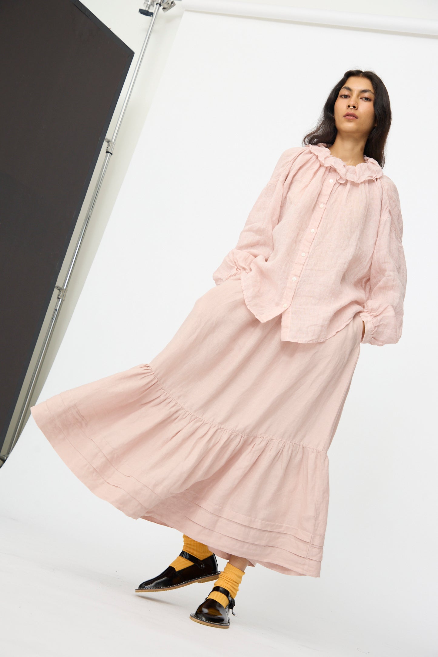 A woman stands in a studio wearing the nest Robe Linen Natural Dyed Pleated Gathered Skirt in Pink, paired with a loose light pink blouse, yellow socks, and black shoes against a white background.