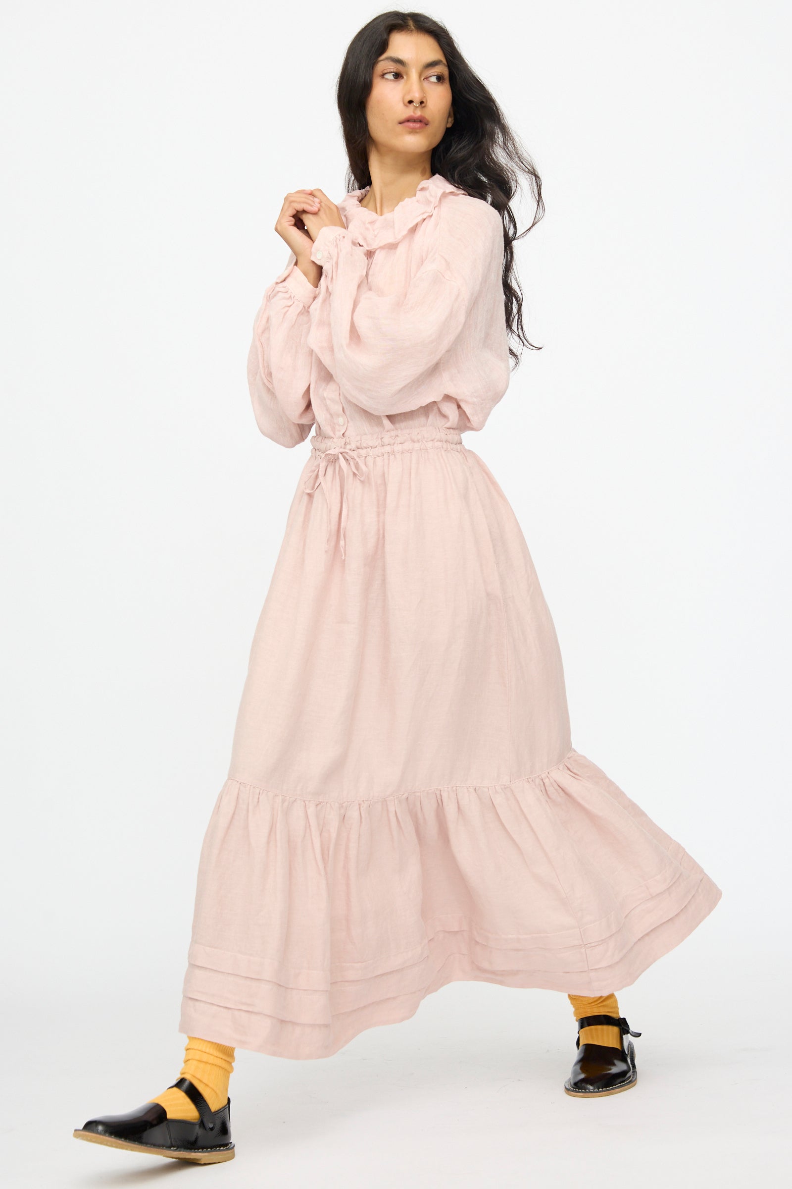 A person wearing the nest Robe Linen Natural Dyed Pleated Gathered Skirt in Pink, paired with yellow socks and black shoes, is walking against a plain white background.