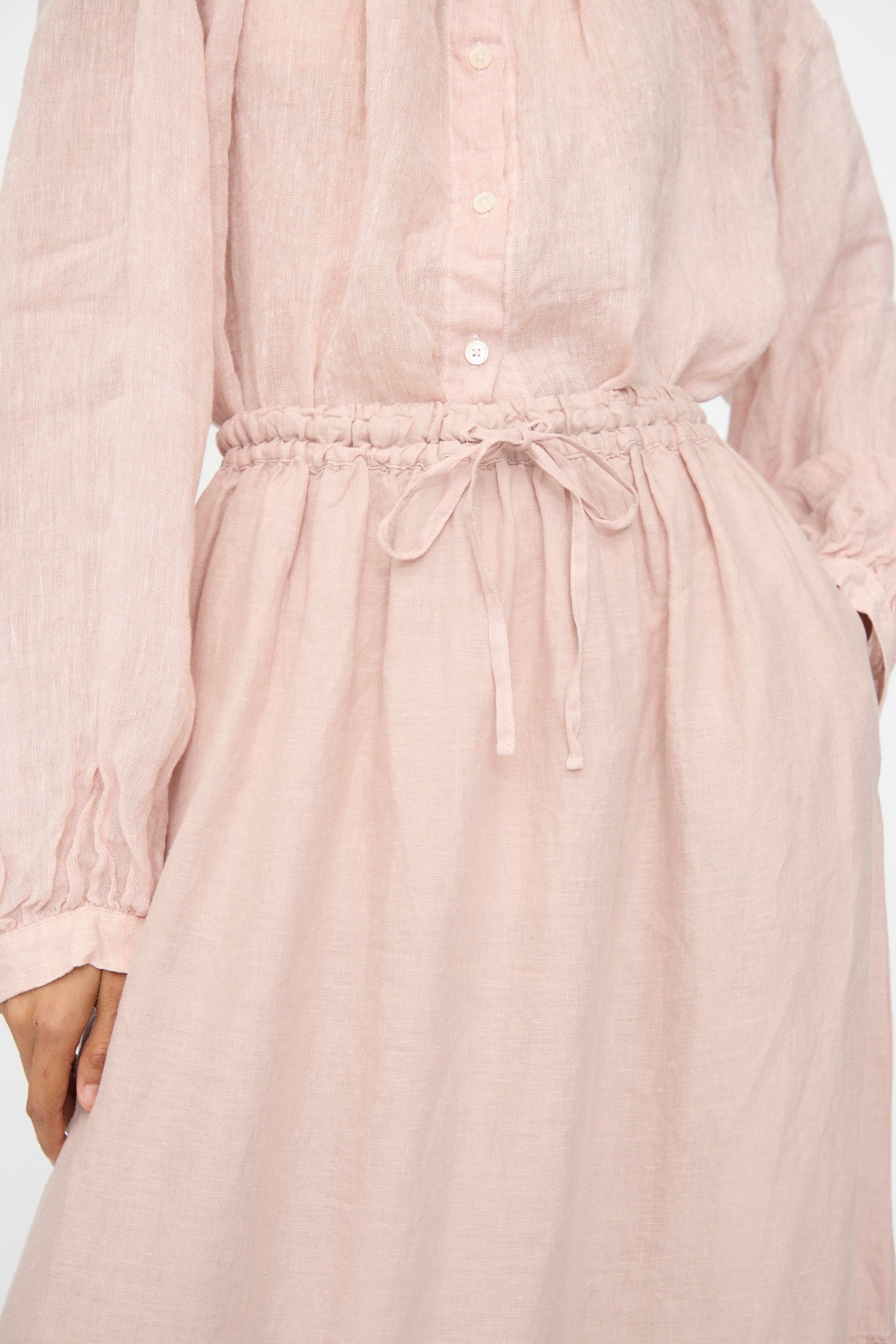 Model wears the nest Robe Linen Natural Dyed Pleated Gathered Skirt in Pink; long-sleeved, button-up dress with gathered waist and drawstring tie layered above, hand partly visible at side.
