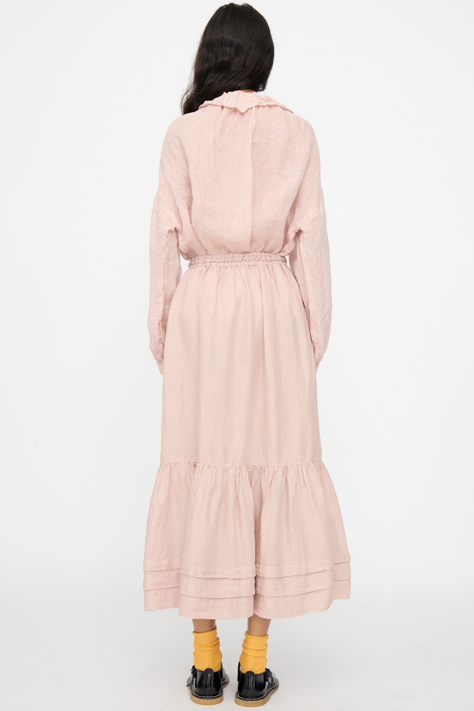 A person stands facing away, wearing the nest Robe Linen Natural Dyed Pleated Gathered Skirt in Pink, a light pink long-sleeve dress with a ruffled hem, yellow socks, and black shoes against a plain white background.