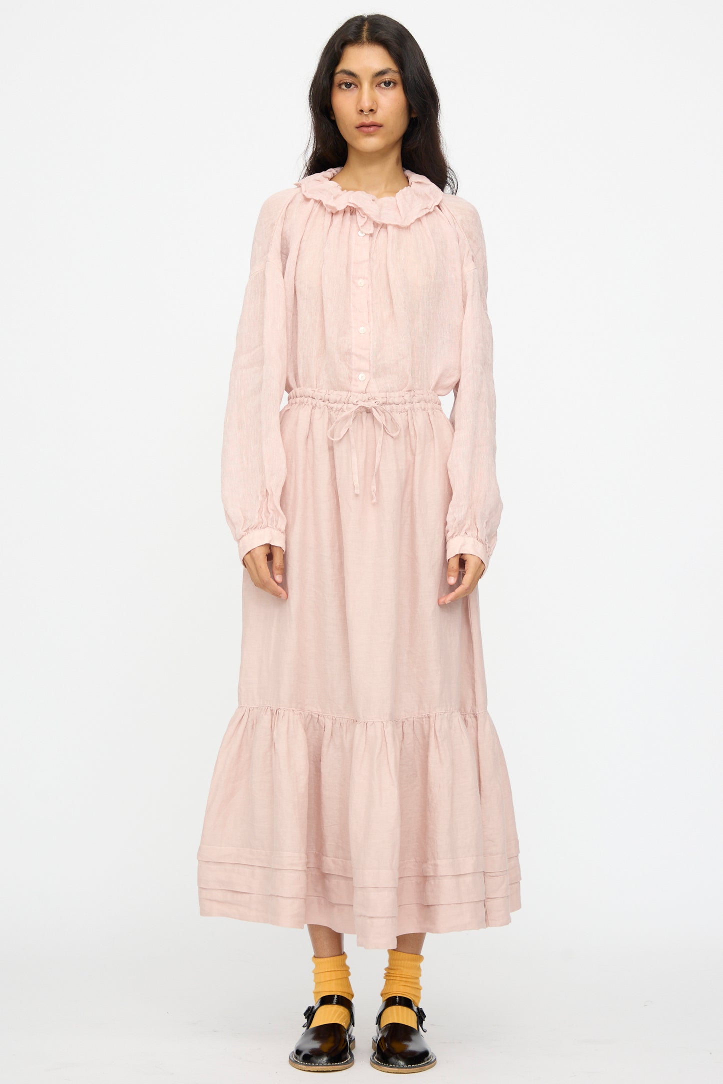 A woman stands against a plain background wearing nest Robe’s Linen Natural Dyed Pleated Gathered Skirt in Pink, styled with a long-sleeved ruffled dress, yellow socks, and black shoes.