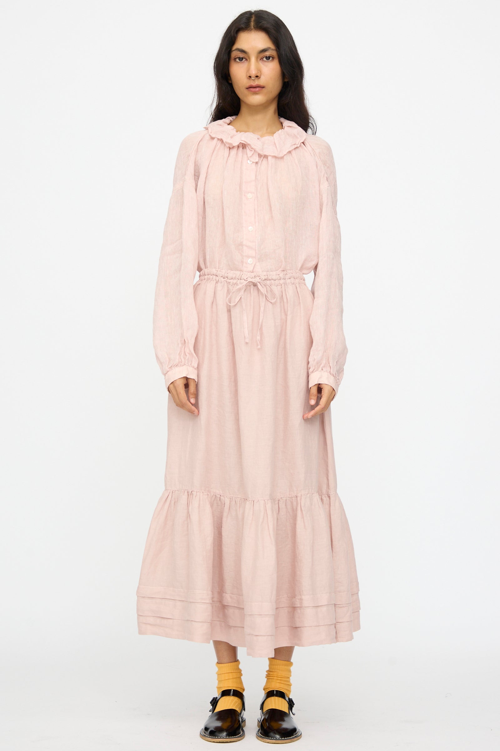 A woman stands against a plain background wearing nest Robe’s Linen Natural Dyed Pleated Gathered Skirt in Pink, styled with a long-sleeved ruffled dress, yellow socks, and black shoes.