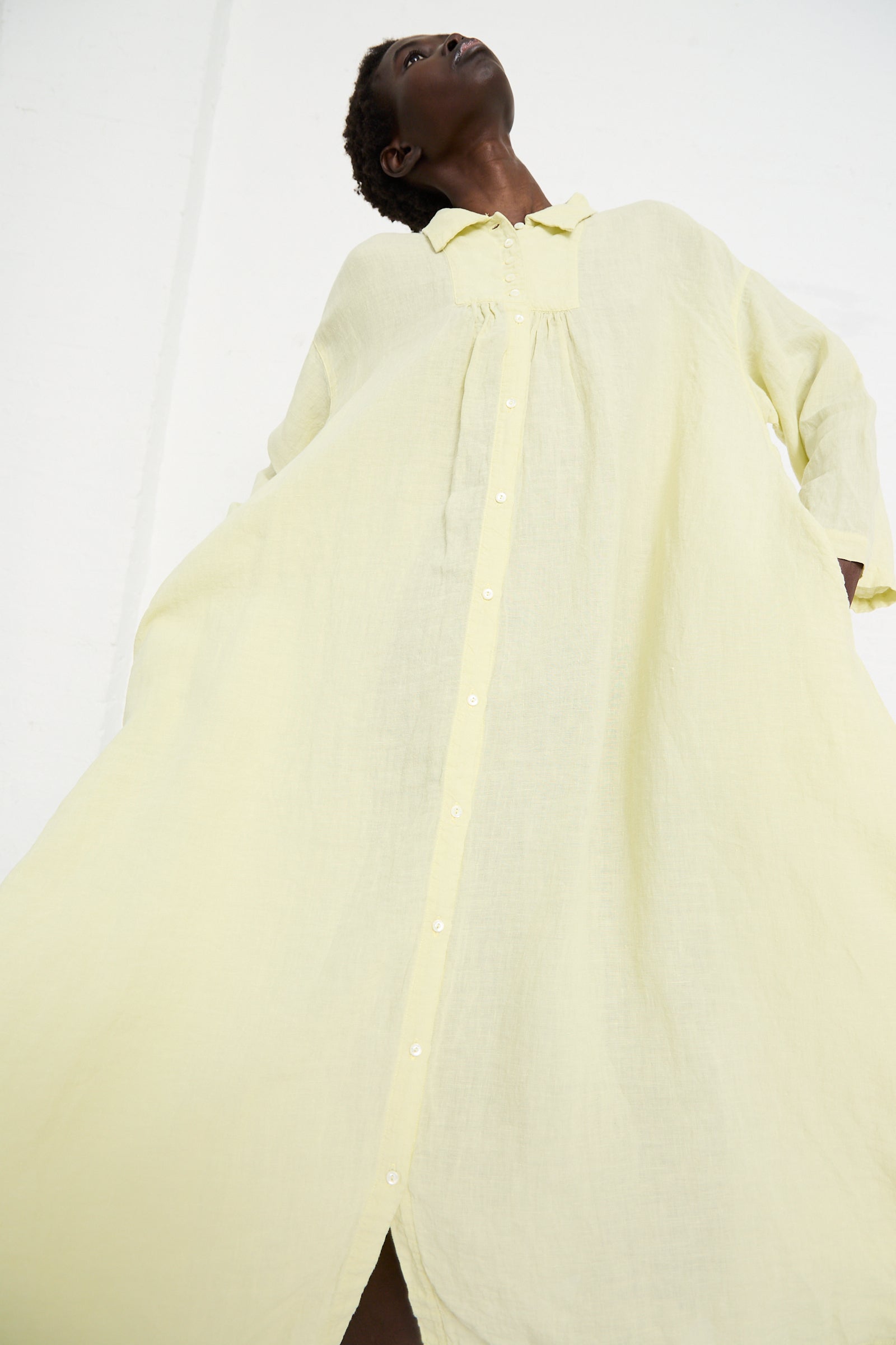 Person wearing a loose, Linen Omi-Zarashi Shirt Dress in Lemon by nest Robe with long sleeves and a button-down front, standing and looking upward. The photo is taken from a low angle against a plain background.