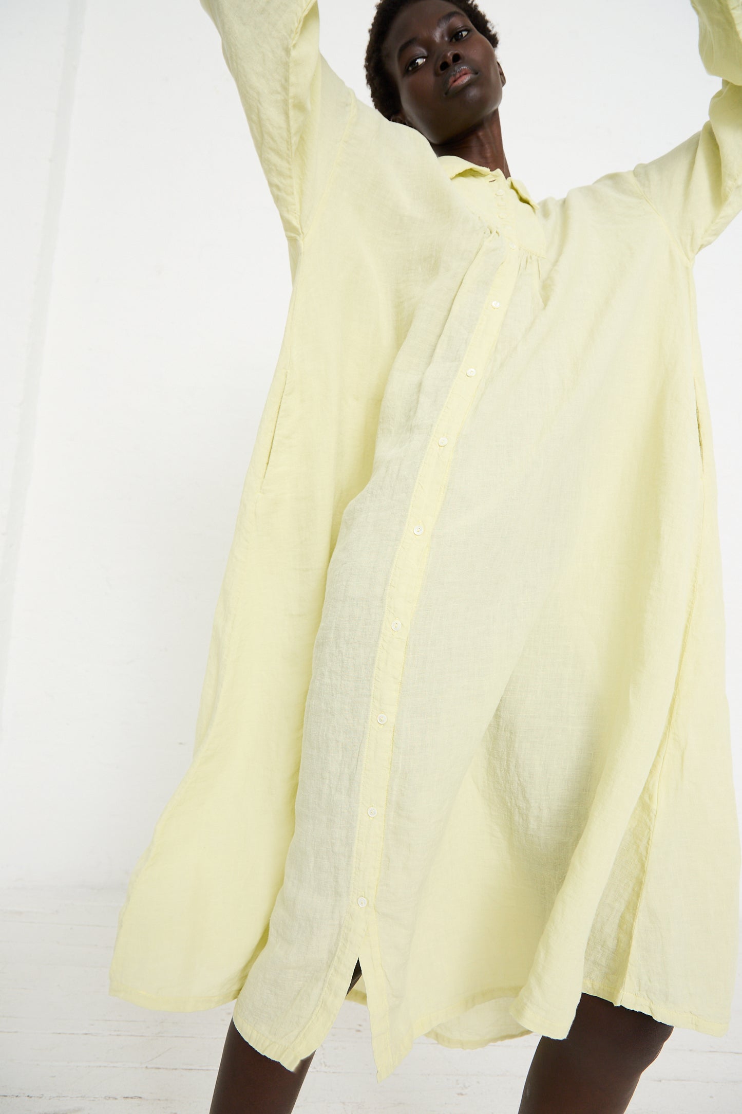 A person wearing a long, Linen Omi-Zarashi Shirt Dress in Lemon by nest Robe with their arms raised, standing against a white background.