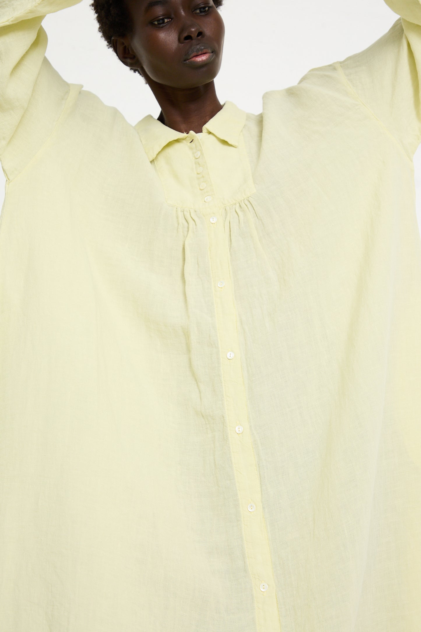 Person wearing a button-up, loose-fitting Linen Omi-Zarashi Shirt Dress in Lemon by nest Robe with arms raised, showcasing the garment's size and textured fabric.