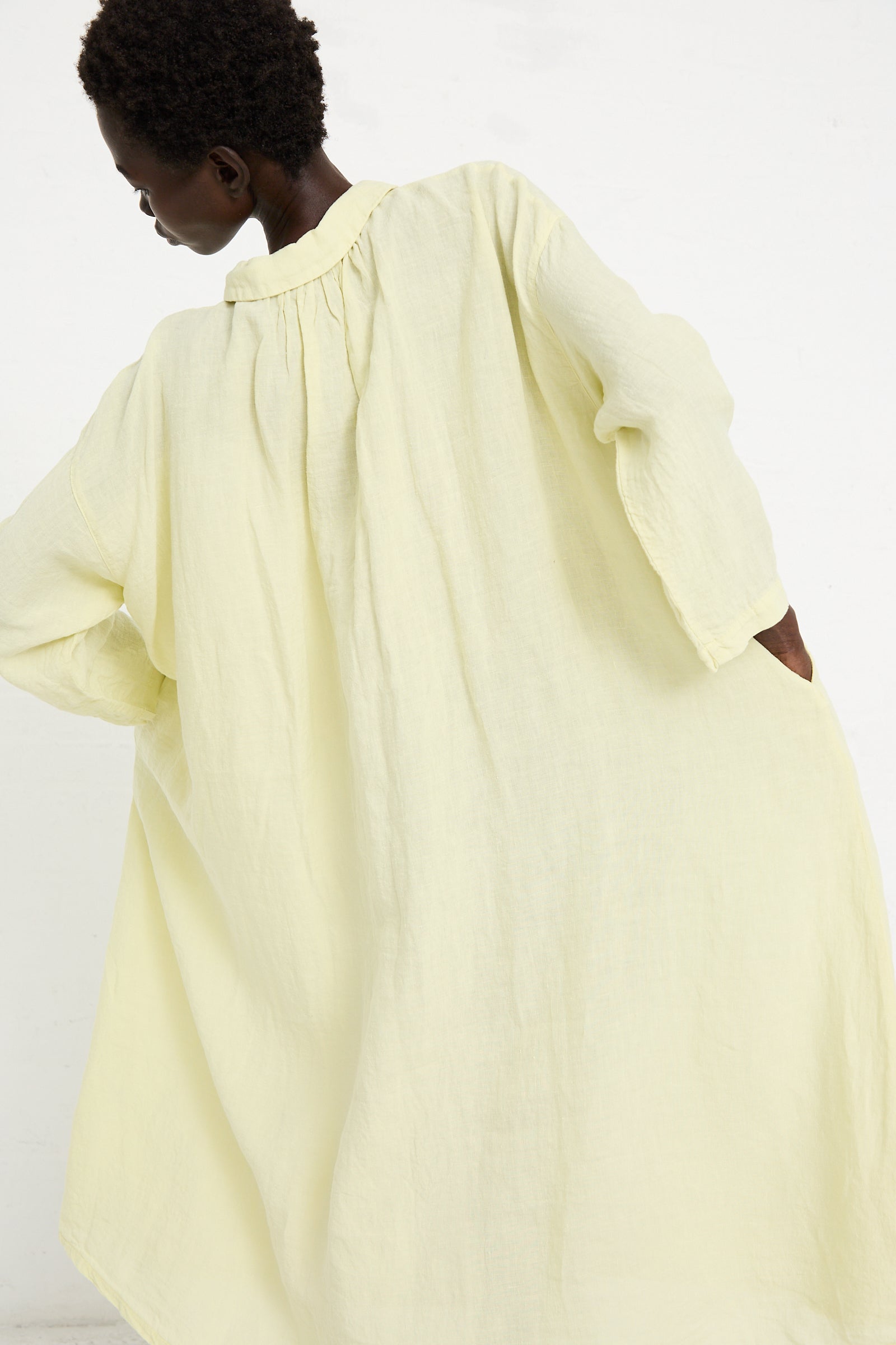 Person wearing a loose-fitting, light yellow textured **Linen Omi-Zarashi Shirt Dress in Lemon** by **nest Robe**, pictured from the back and slightly turned to the side, standing against a plain white background.