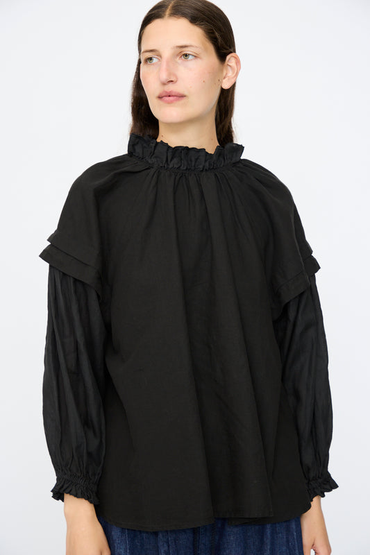 A person models the nest Robe Linen Ramie Frilled Two-Way Blouse in Black, featuring long sleeves and a ruffled neck, against a plain white background.