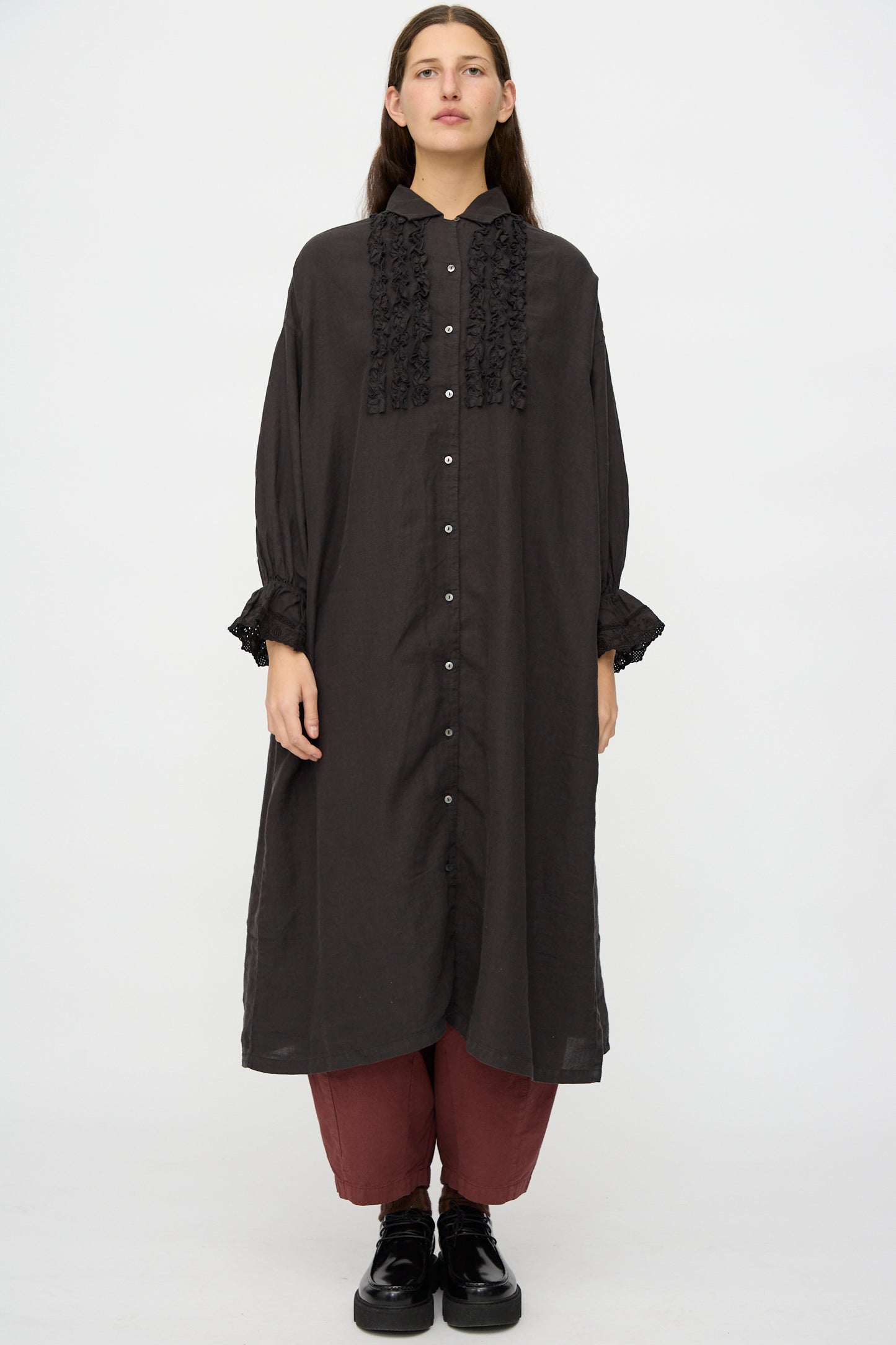 A person wearing the nest Robe Linen Scallop Lace Dress in Ink Black over maroon pants and black shoes, standing against a plain white background.