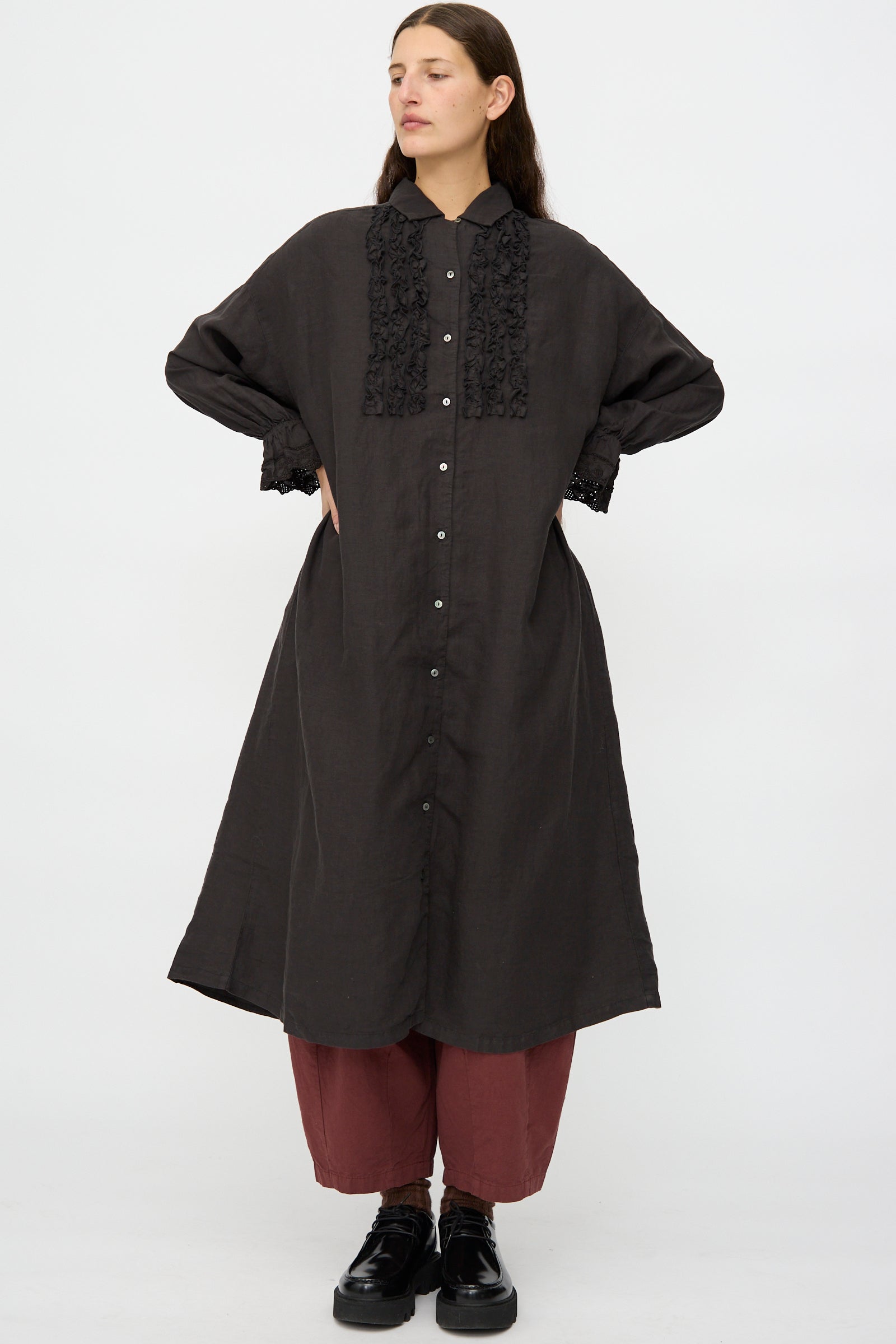 A person faces forward against a white background wearing the nest Robe Linen Scallop Lace Dress in Ink Black, paired with maroon pants and black shoes.