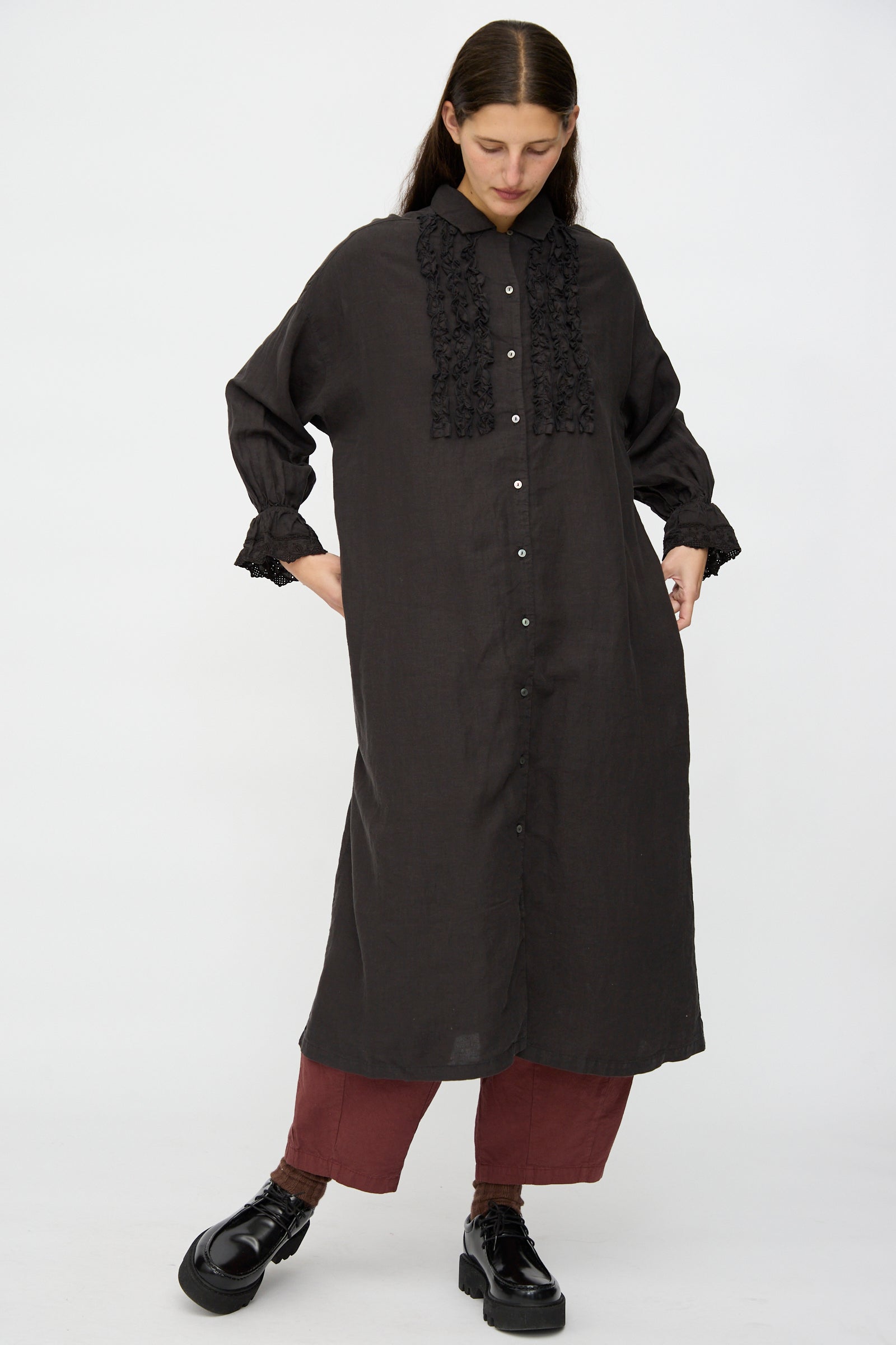 A person wears the nest Robe Linen Scallop Lace Dress in Ink Black with maroon pants and black shoes, standing against a plain white background.
