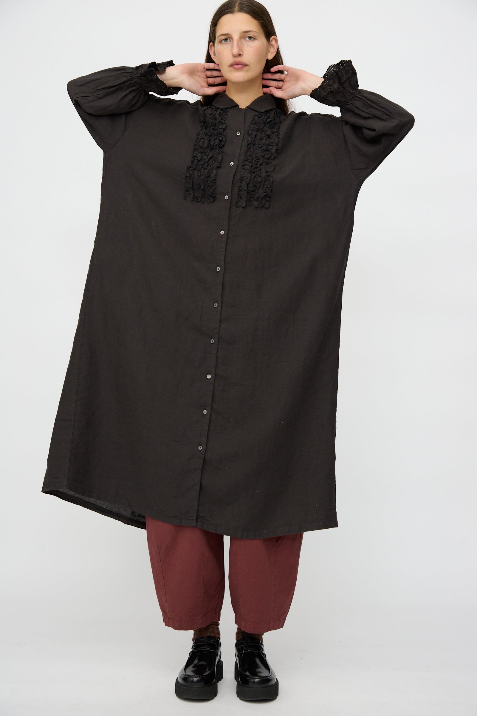 Model wearing the nest Robe Linen Scallop Lace Dress in Ink Black, featuring loose long sleeves and ruffle details, styled with red pants and black shoes against a plain background.