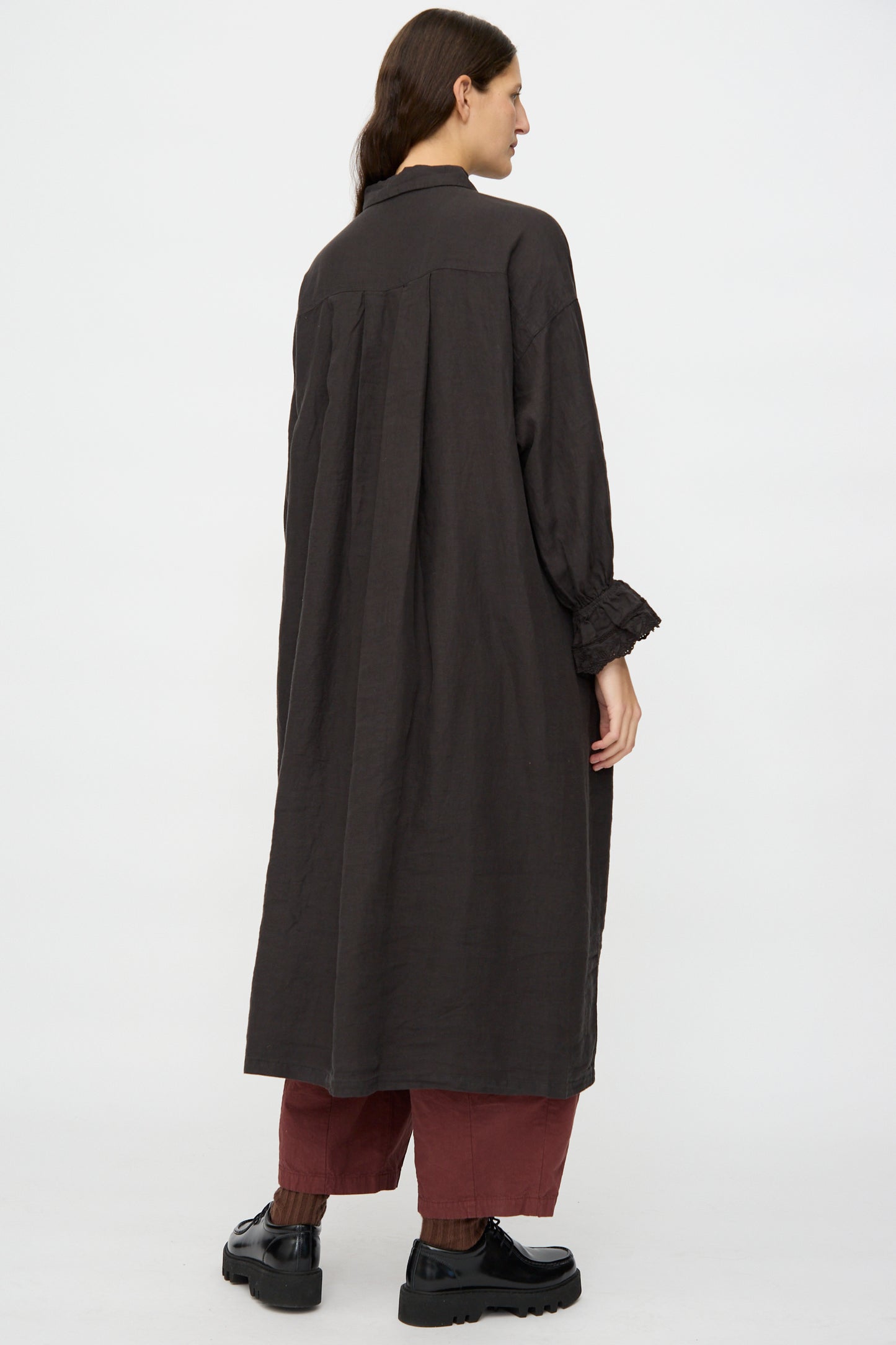 A person stands facing away against a plain light background, wearing nest Robe's Linen Scallop Lace Dress in Ink Black over red pants and black shoes.