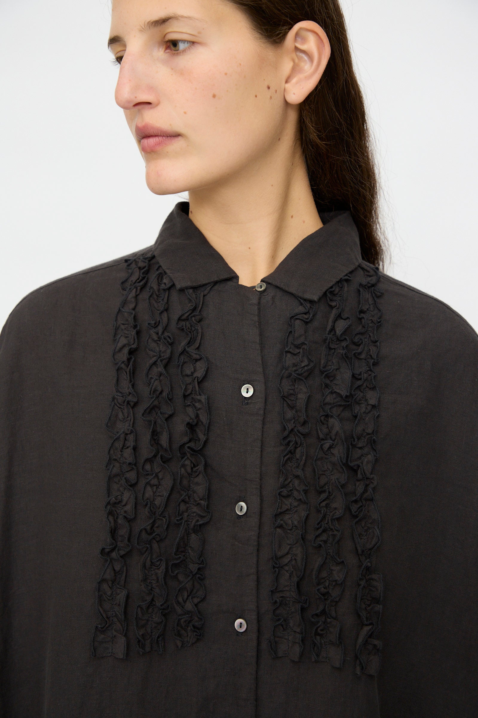 A person, shown from the shoulders up against a plain background, wears the nest Robe Linen Scallop Lace Dress in Ink Black, featuring vertical ruffle details on the front.