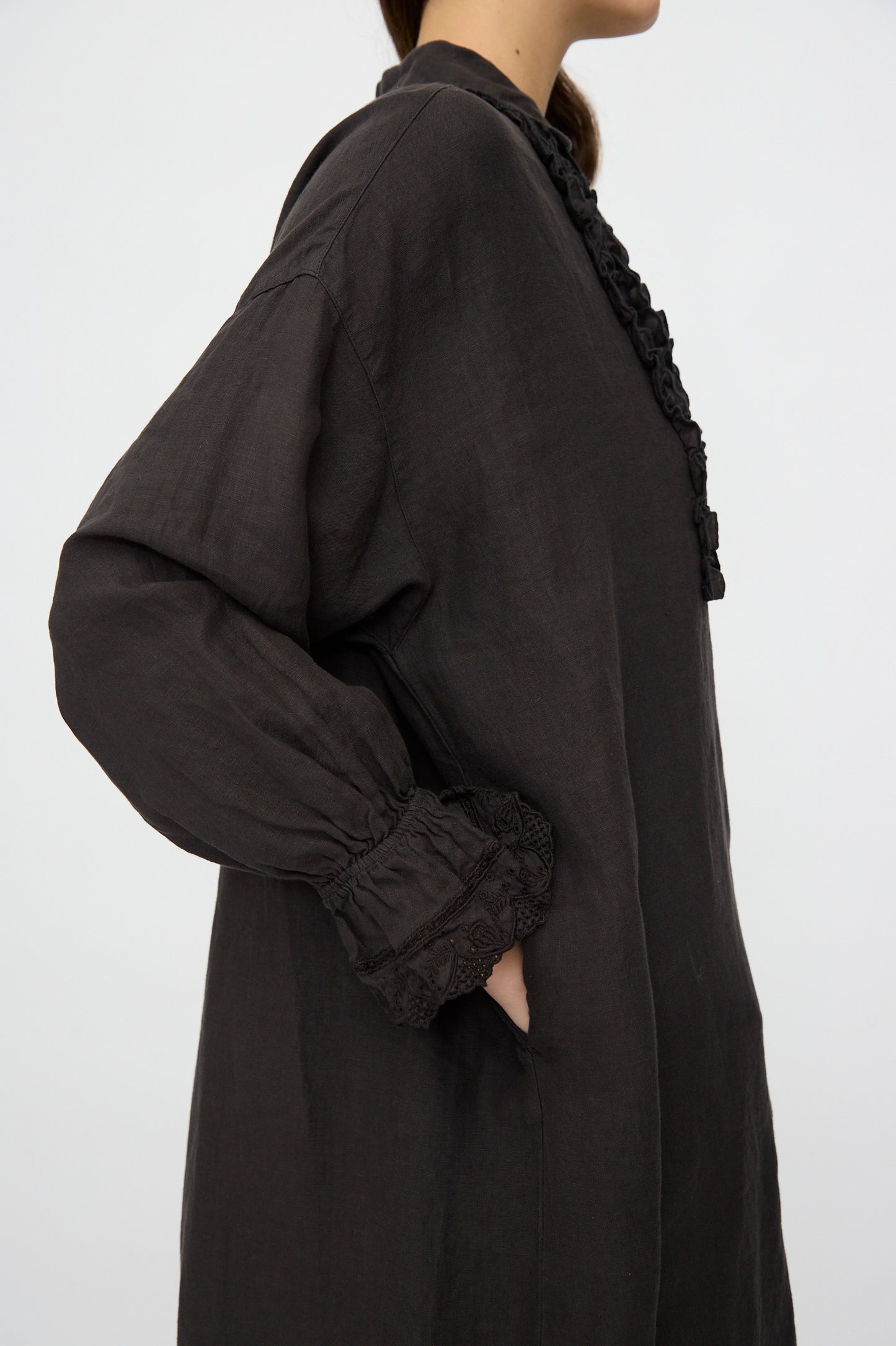 A person wearing the nest Robe Linen Scallop Lace Dress in Ink Black stands sideways with one hand in the pocket against a plain background.