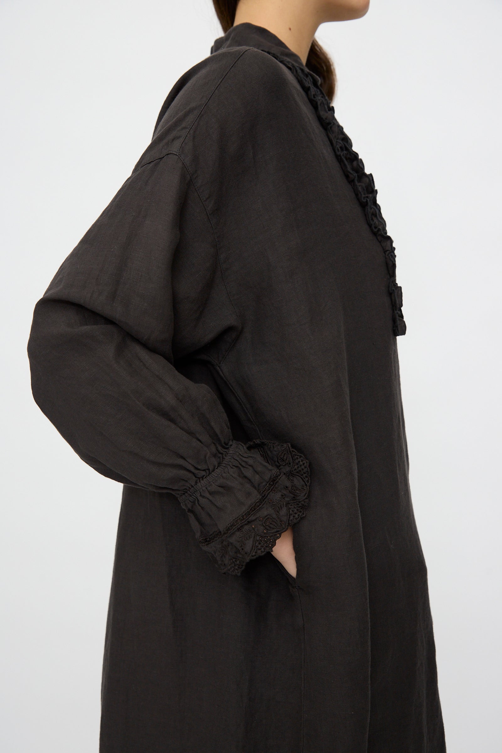 A person wearing the nest Robe Linen Scallop Lace Dress in Ink Black stands sideways with one hand in the pocket against a plain background.