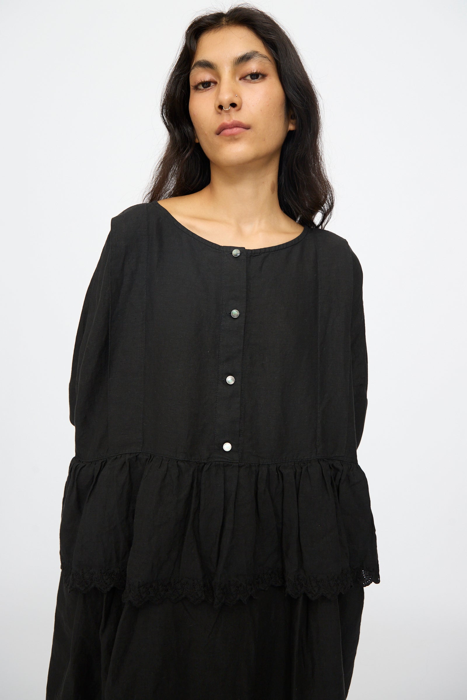 A woman with long dark hair wears the nest Robe Linen Scallop Lace Two-Way Peplum Dress in Black, featuring a loose fit and button front, standing against a plain white background.