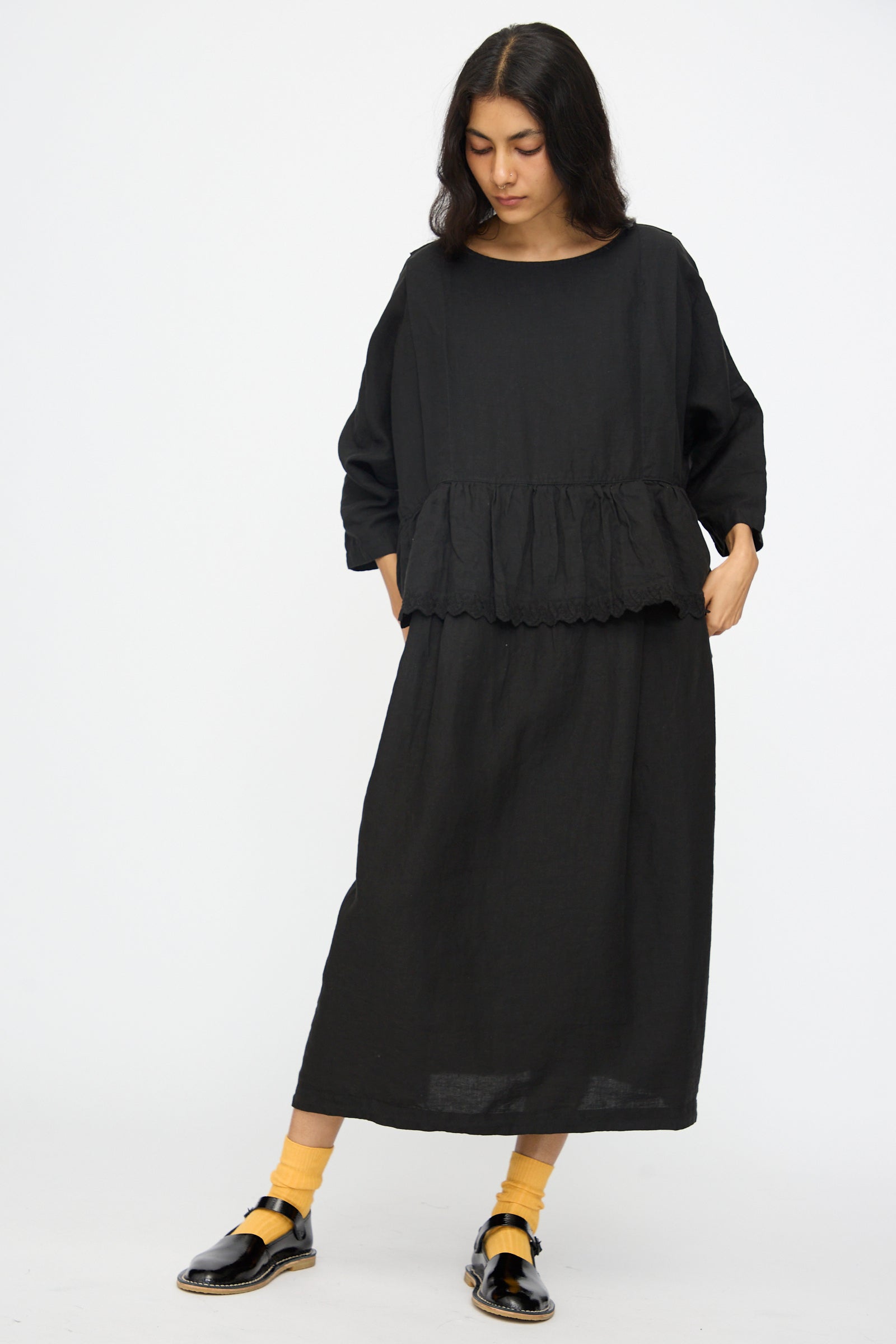 A woman poses against a white background in nest Robe’s Linen Scallop Lace Two-Way Peplum Dress in black, paired with yellow socks and black shoes. She stands with hands in pockets, head tilted. Made in Japan, this look is effortlessly chic.