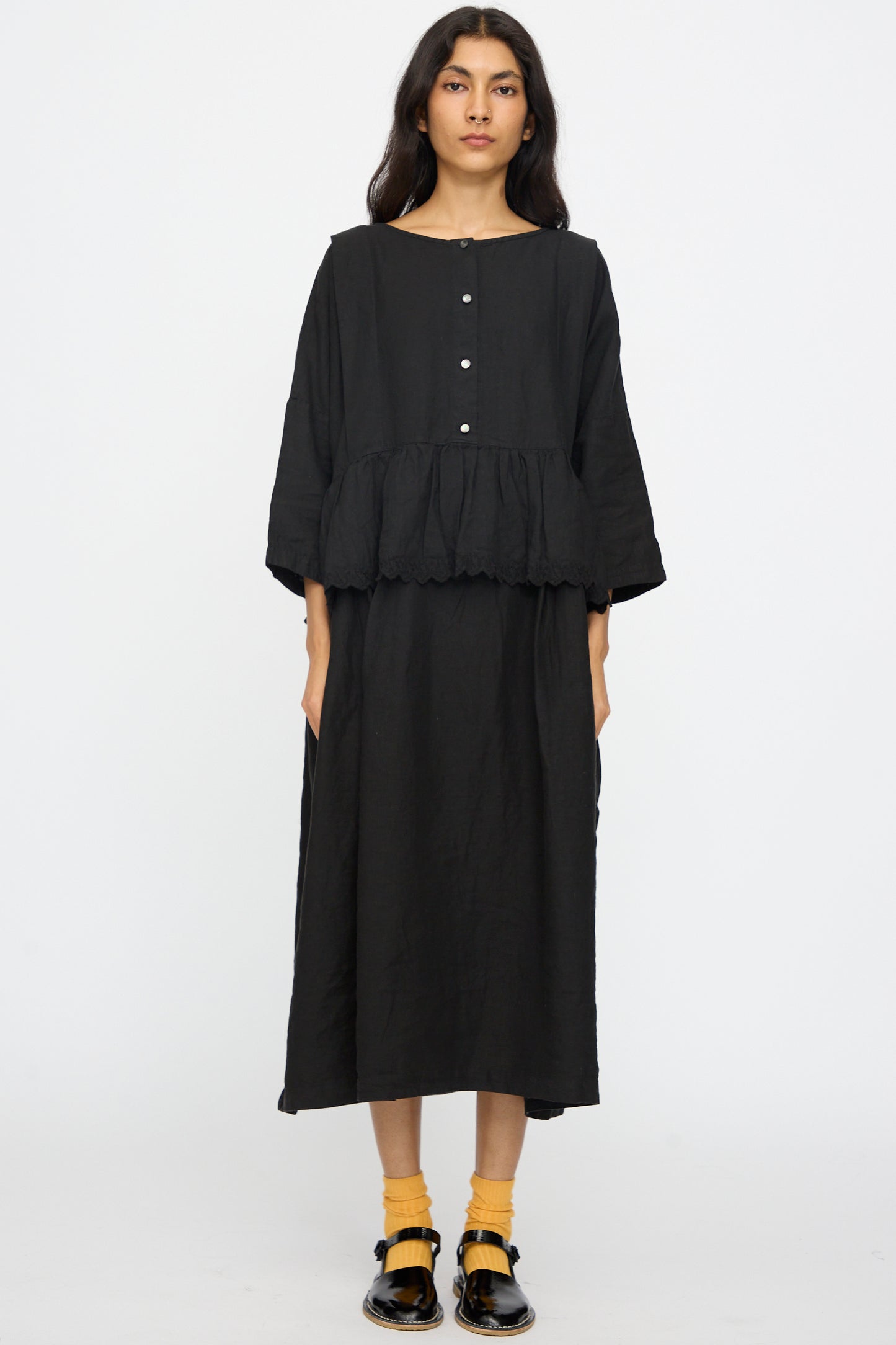 A person stands against a plain background wearing the nest Robe Linen Scallop Lace Two-Way Peplum Dress in Black, styled with yellow socks and black shoes.