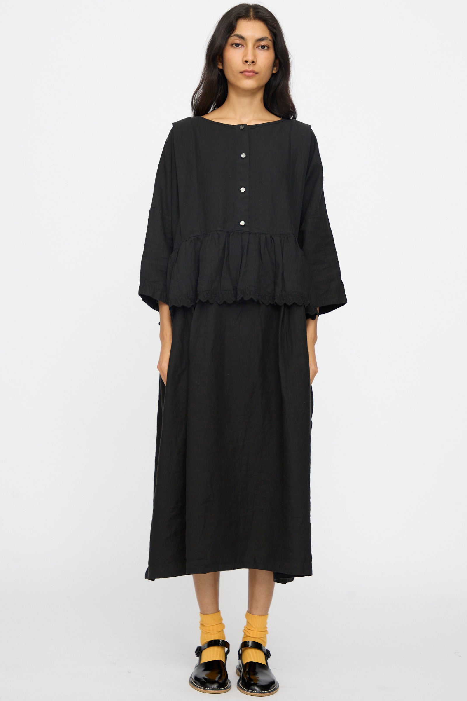 A person stands against a plain background wearing the nest Robe Linen Scallop Lace Two-Way Peplum Dress in Black, styled with yellow socks and black shoes.
