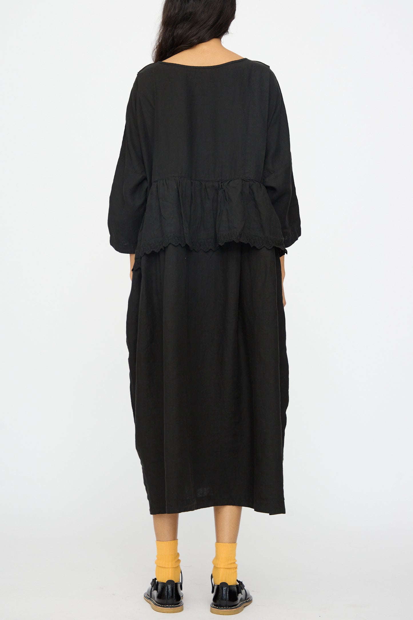 A person wears the nest Robe Linen Scallop Lace Two-Way Peplum Dress in black, paired with yellow socks and black shoes, photographed from behind against a white background. This reversible Japanese-made dress features simple, quality craftsmanship.