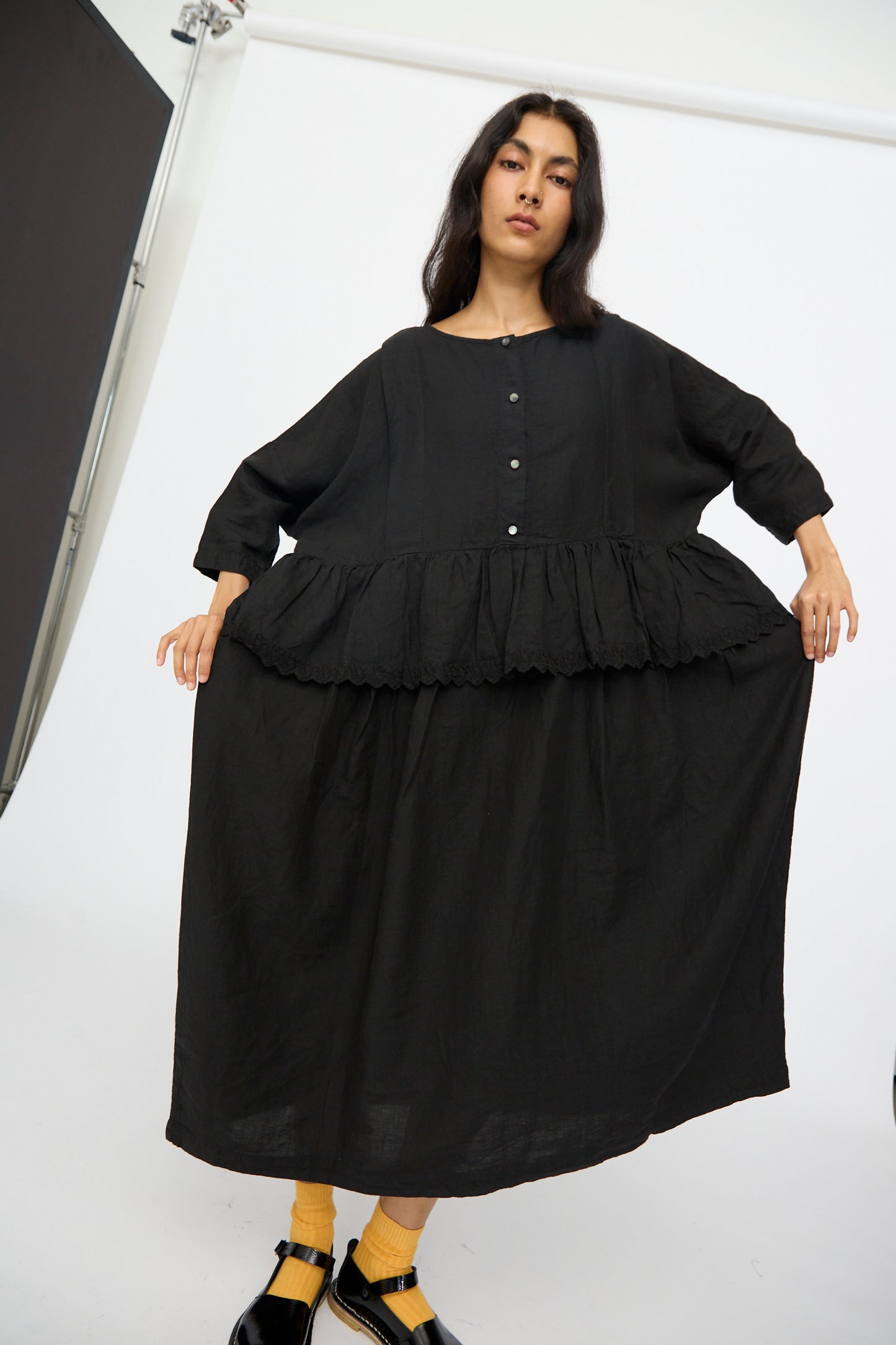 A woman in the nest Robe Linen Scallop Lace Two-Way Peplum Dress in Black stands in a studio, holding out the skirt. This elegant, loose-fitting dress is crafted in Japan.