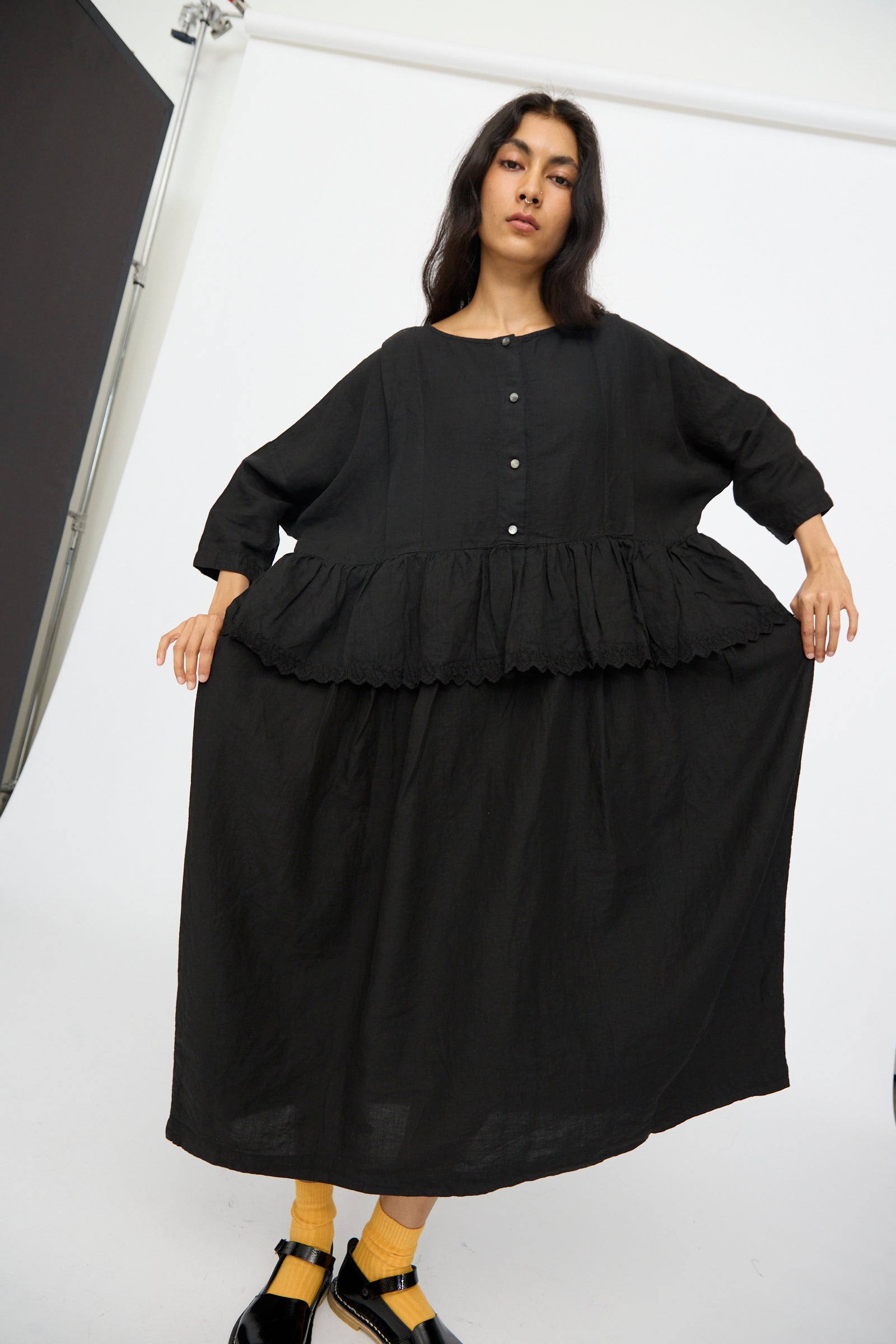A woman in the nest Robe Linen Scallop Lace Two-Way Peplum Dress in Black stands in a studio, holding out the skirt. This elegant, loose-fitting dress is crafted in Japan.
