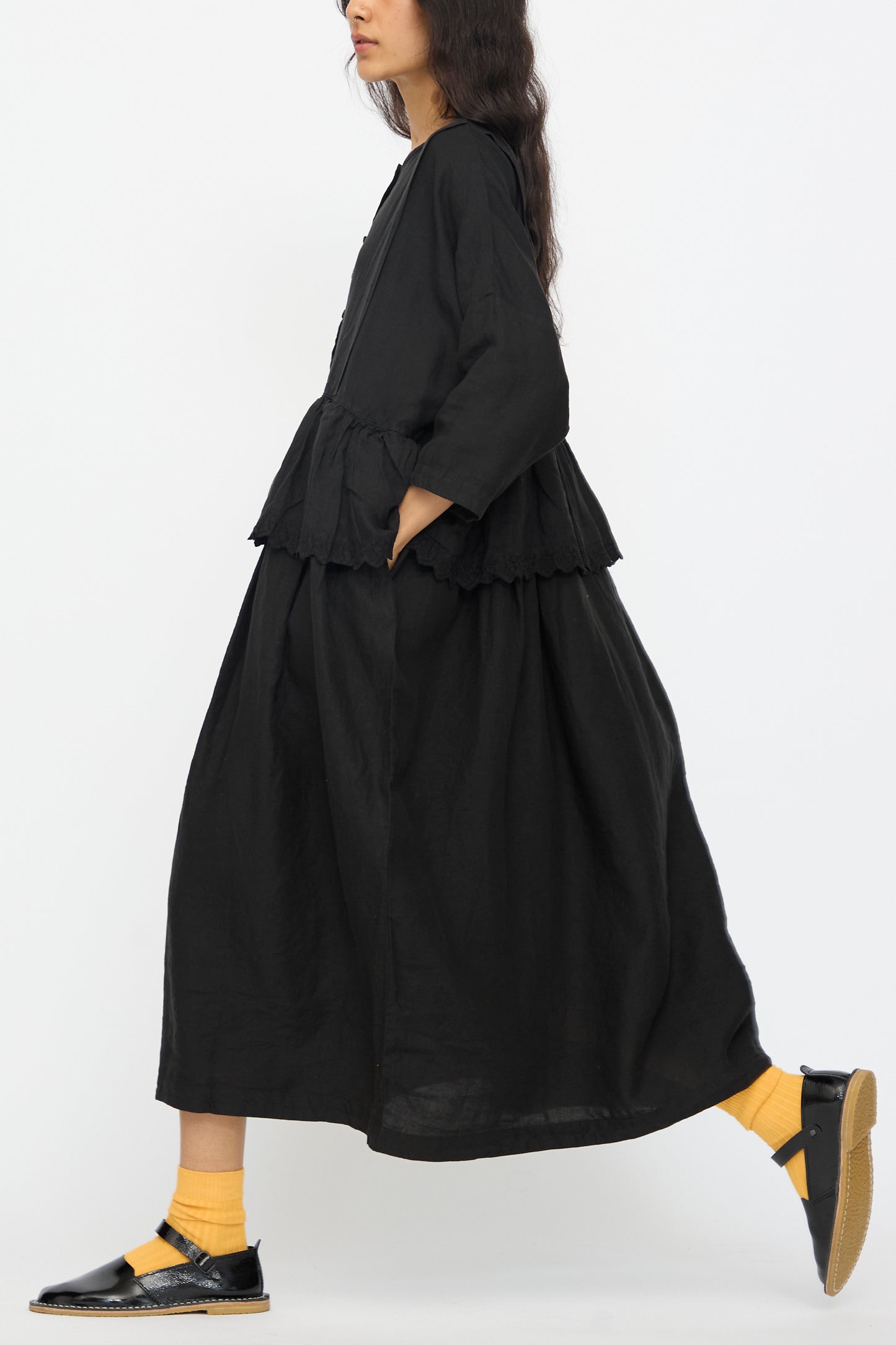 A woman is walking in a nest Robe Linen Scallop Lace Two-Way Peplum Dress in black, styled with yellow socks and black sandals, photographed in profile against a plain white background.