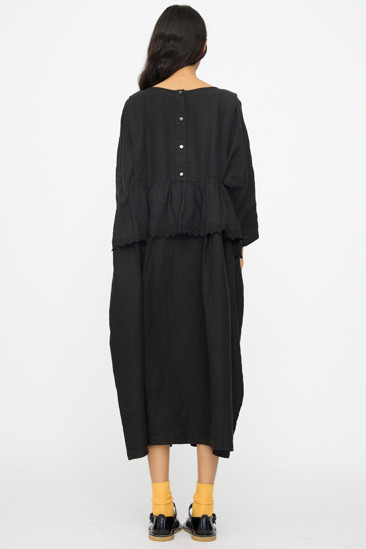 A person with long dark hair, wearing the nest Robe Linen Scallop Lace Two-Way Peplum Dress in Black—made in Japan—with yellow socks and black shoes, stands facing away from the camera against a plain white background.