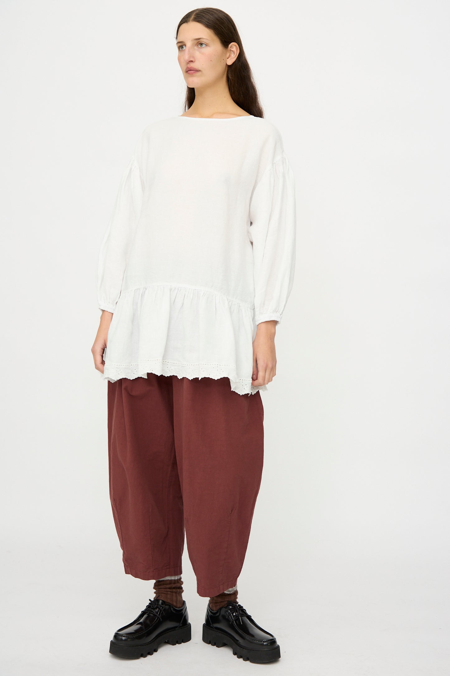 A person stands against a plain background wearing the nest Robe Linen Scallop Lace Two-Way Tunic in white with bishop sleeves, wide maroon pants, brown socks, and black shoes.