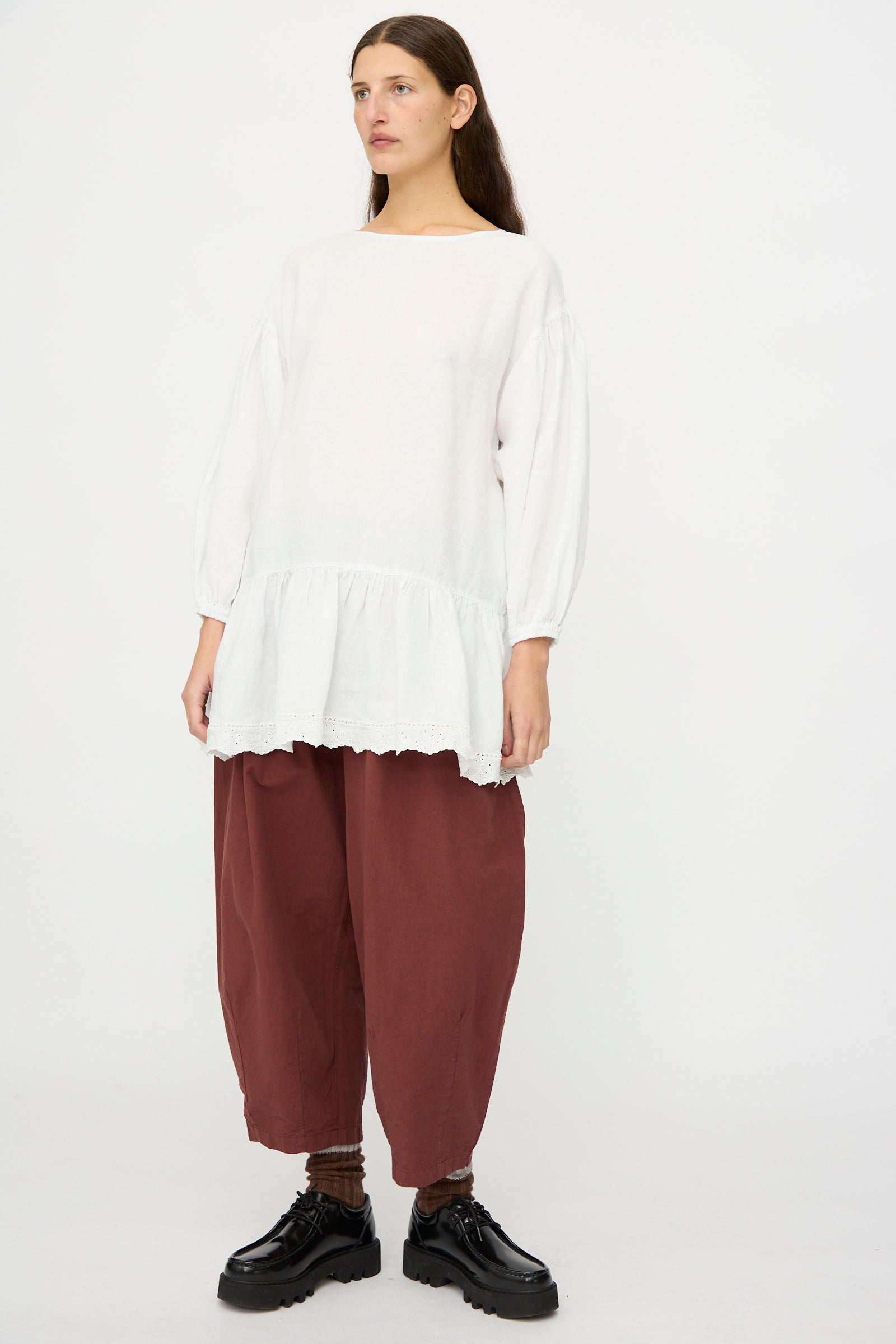 A person stands against a plain background wearing the nest Robe Linen Scallop Lace Two-Way Tunic in white with bishop sleeves, wide maroon pants, brown socks, and black shoes.