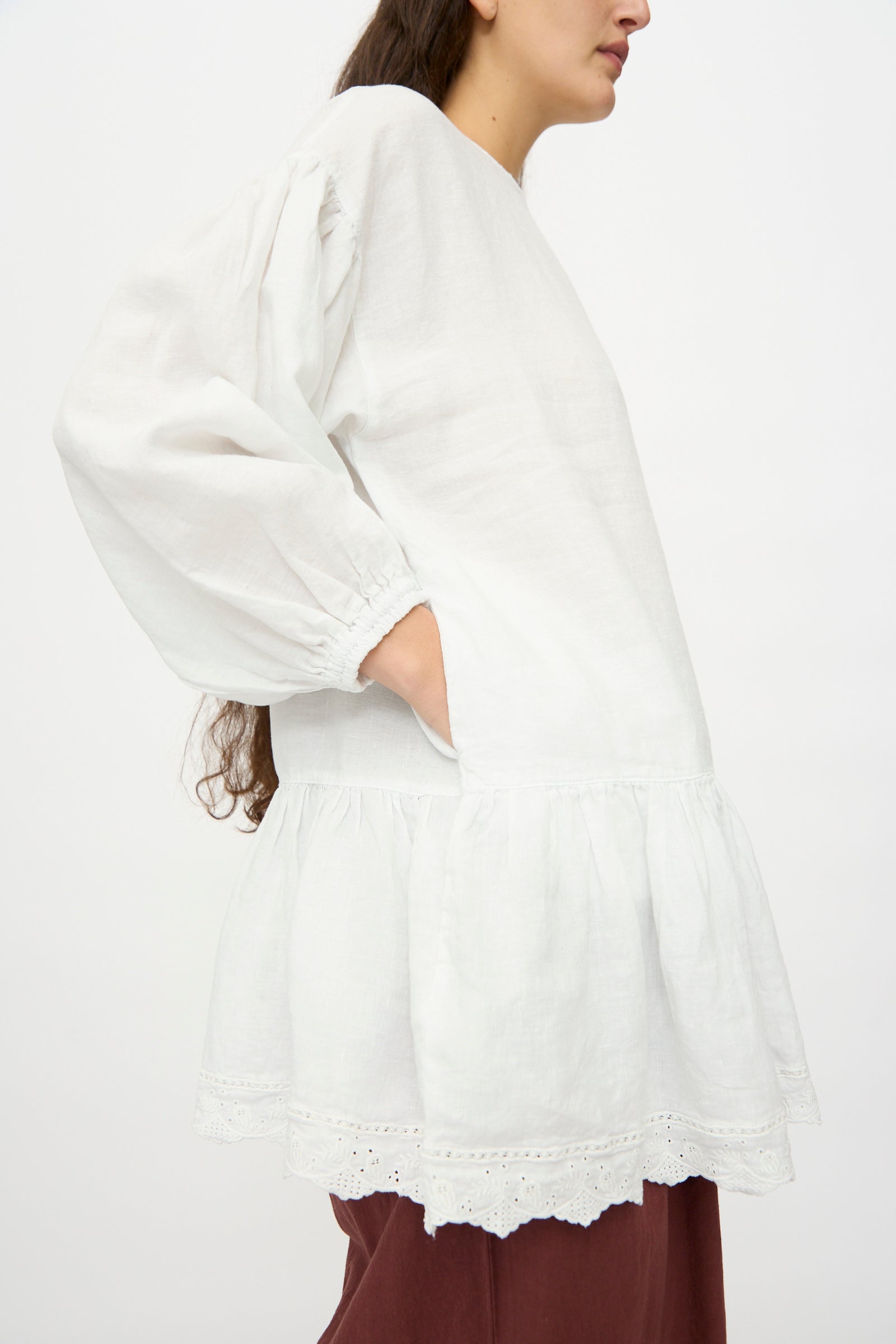 A model in the nest Robe Linen Scallop Lace Two-Way Tunic, featuring a loose fit with bishop sleeves and lace embroidery, is paired with maroon pants and stands sideways with one hand in a pocket.