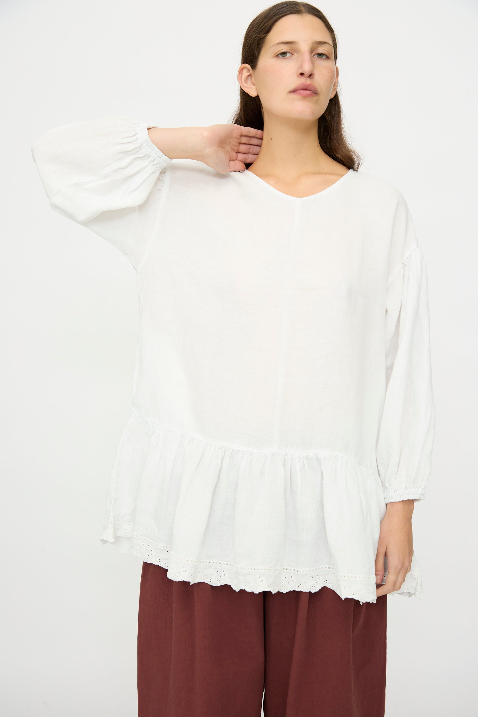 A person poses against a plain background wearing nest Robe's Linen Scallop Lace Two-Way Tunic—a loose-fitting white blouse with delicate lace embroidery—paired with wide-legged maroon pants, one hand raised to their neck.