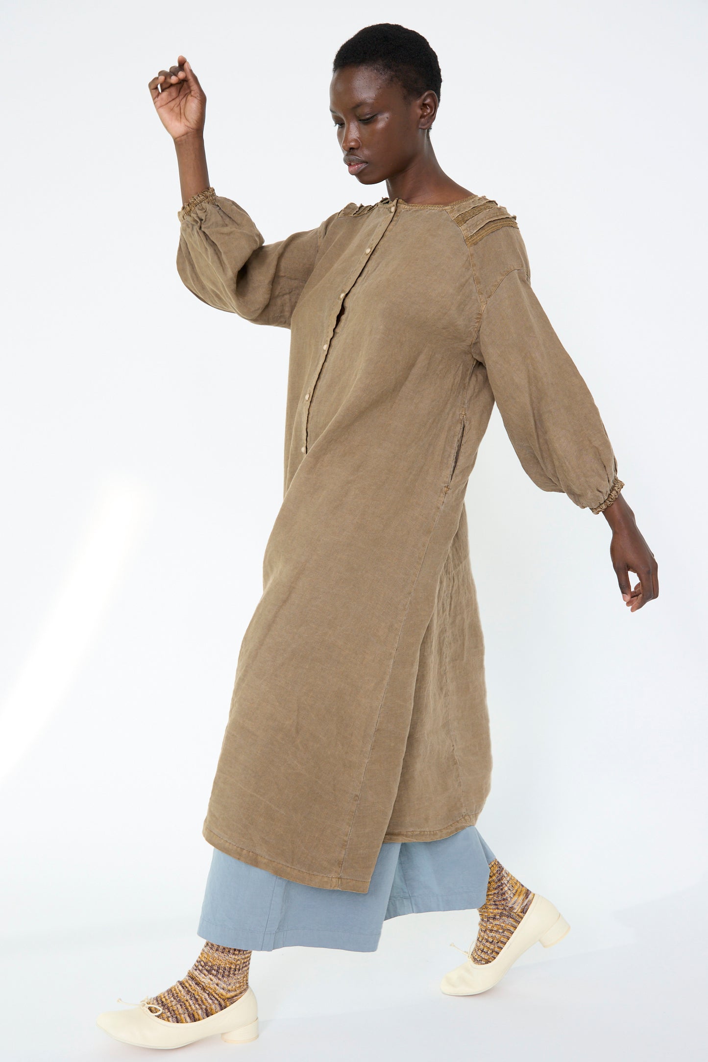 A person walks against a plain background, wearing the nest Robe Natural Dyed Linen Cotton Lace Dress in camel over light blue pants, paired with patterned socks and white shoes.