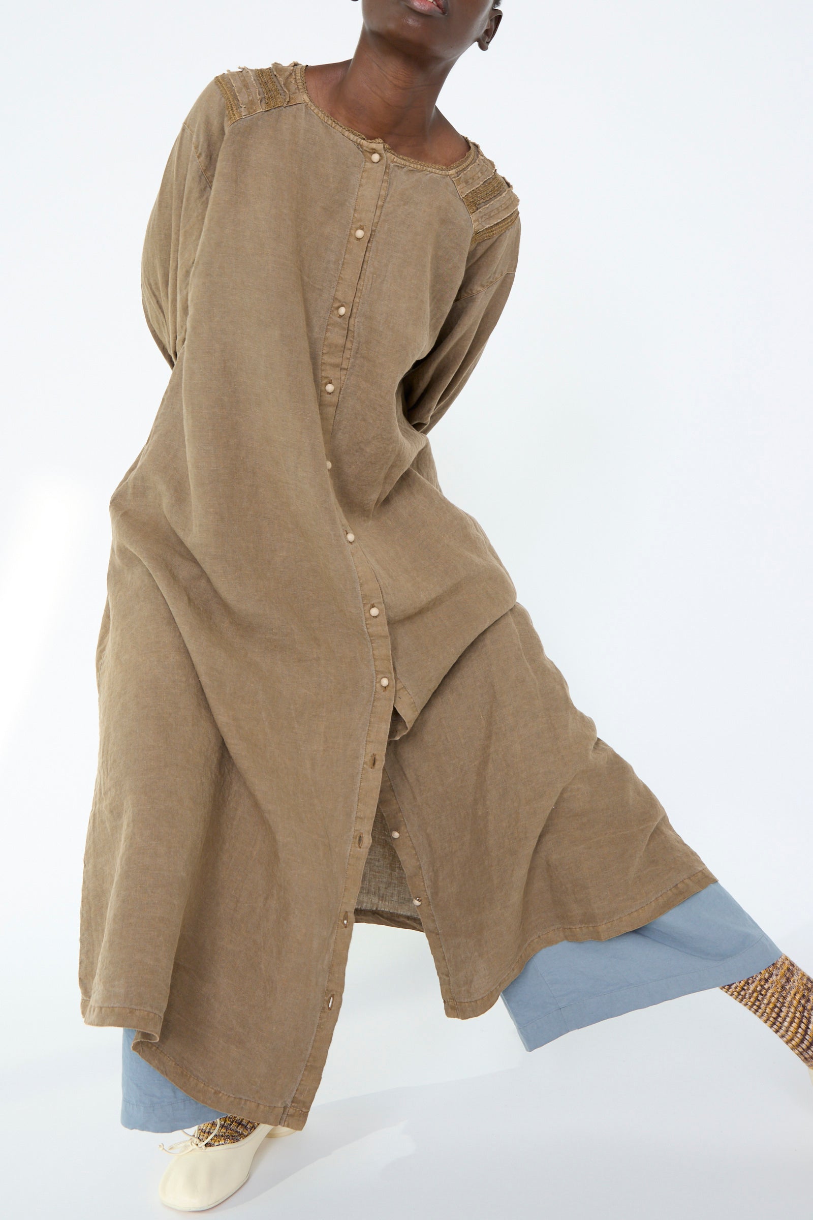 A person wearing the Nest Robe Natural Dyed Linen Cotton Lace Dress in Camel, featuring intricate shoulder detailing, paired with light blue pants against a white background.