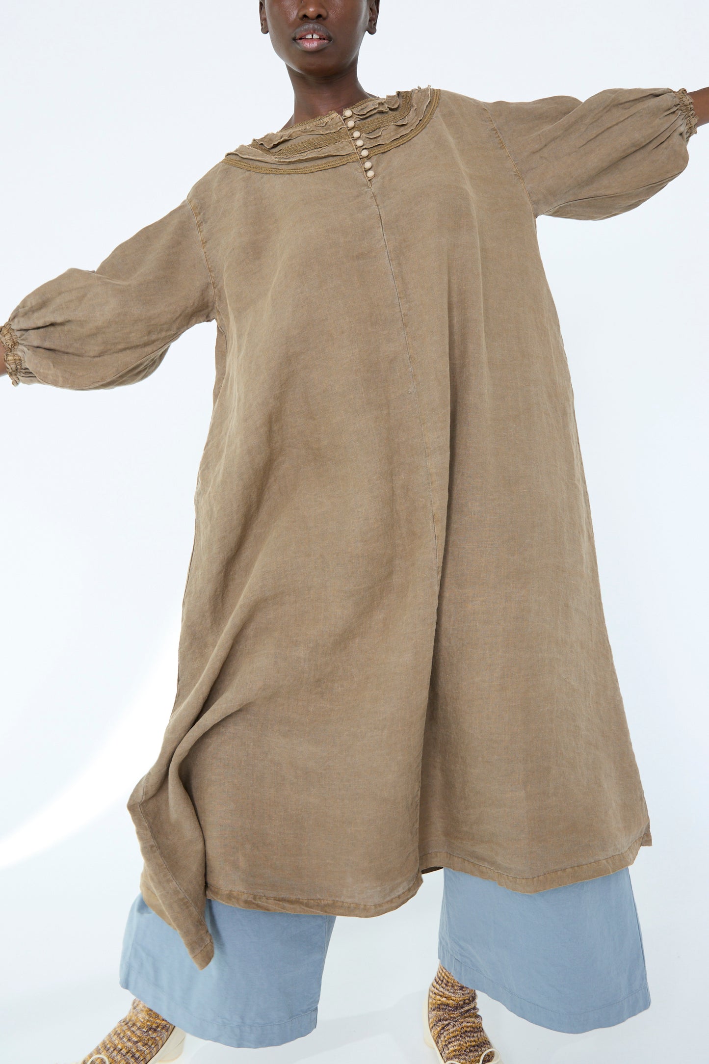 A person stands against a white background with outstretched arms, wearing the naturally dyed camel-colored linen cotton lace dress from nest Robe over light blue pants.