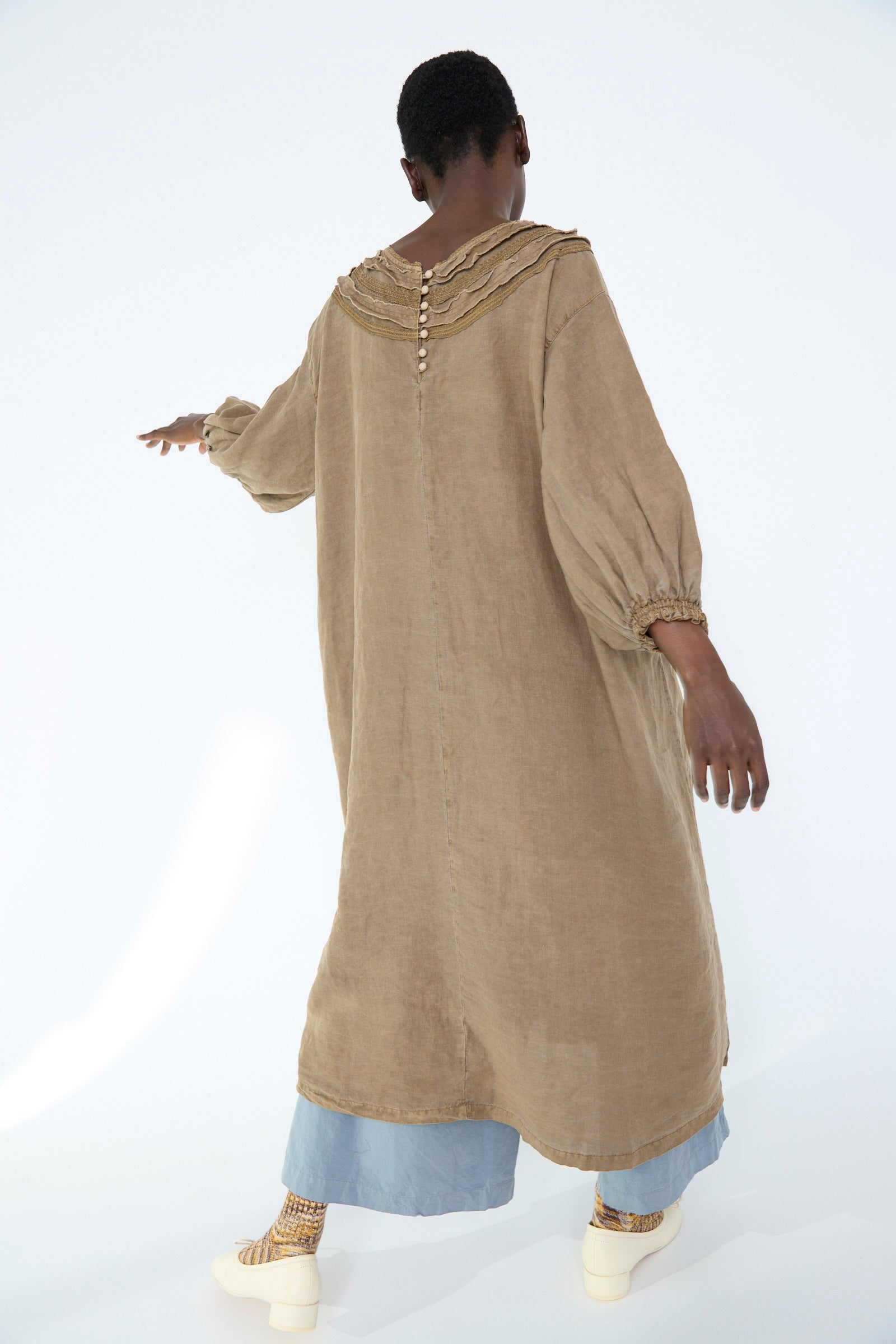 A person wearing the nest Robe Natural Dyed Linen Cotton Lace Dress in Camel, layered over light blue pants against a white background. 