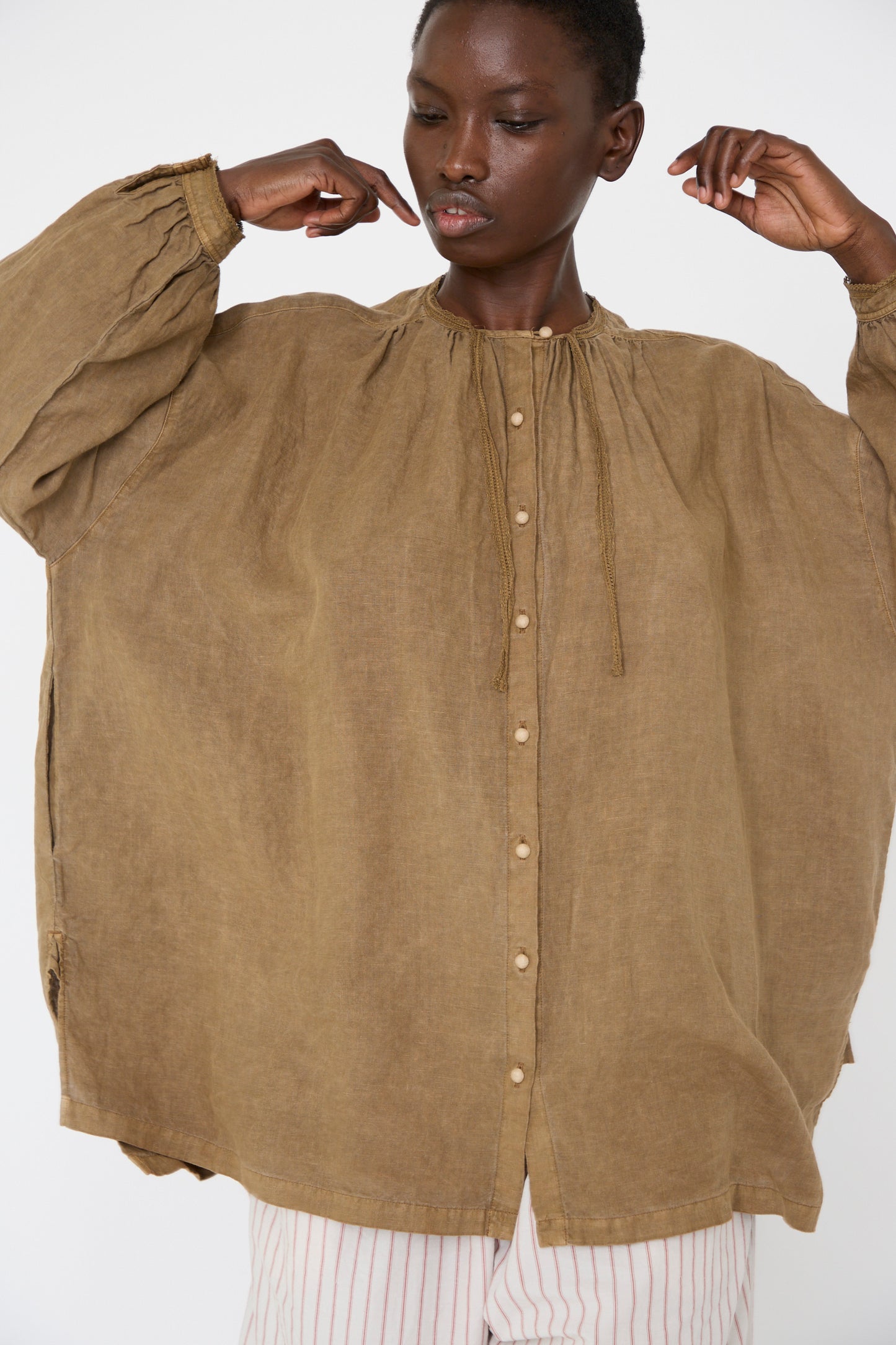 Against a plain background, a woman is wearing the Natural Dyed Linen Cotton Lace Shirt in Camel by nest Robe, paired with off-white striped pants.