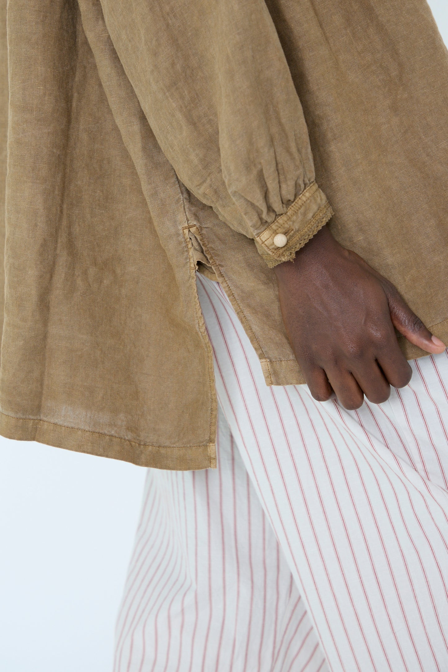 A close up of a woman wearing a Natural Dyed Linen Cotton Lace Shirt in Camel from nest Robe, along with white pants featuring red stripes, with her hand resting on her side.