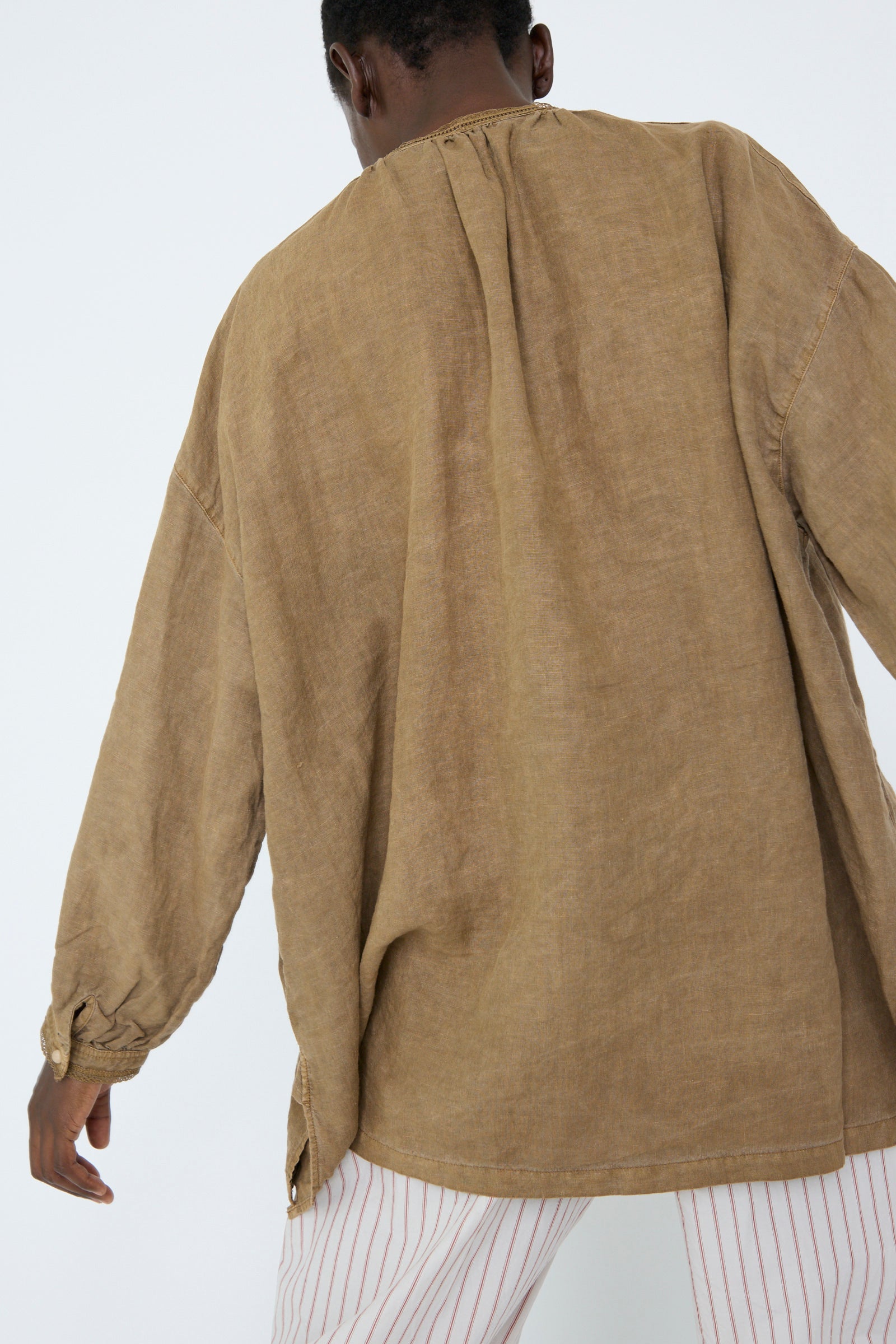 A woman dressed in a loose, oversized Natural Dyed Linen Cotton Lace Shirt in Camel by nest Robe, paired with striped pants, standing with her back turned.