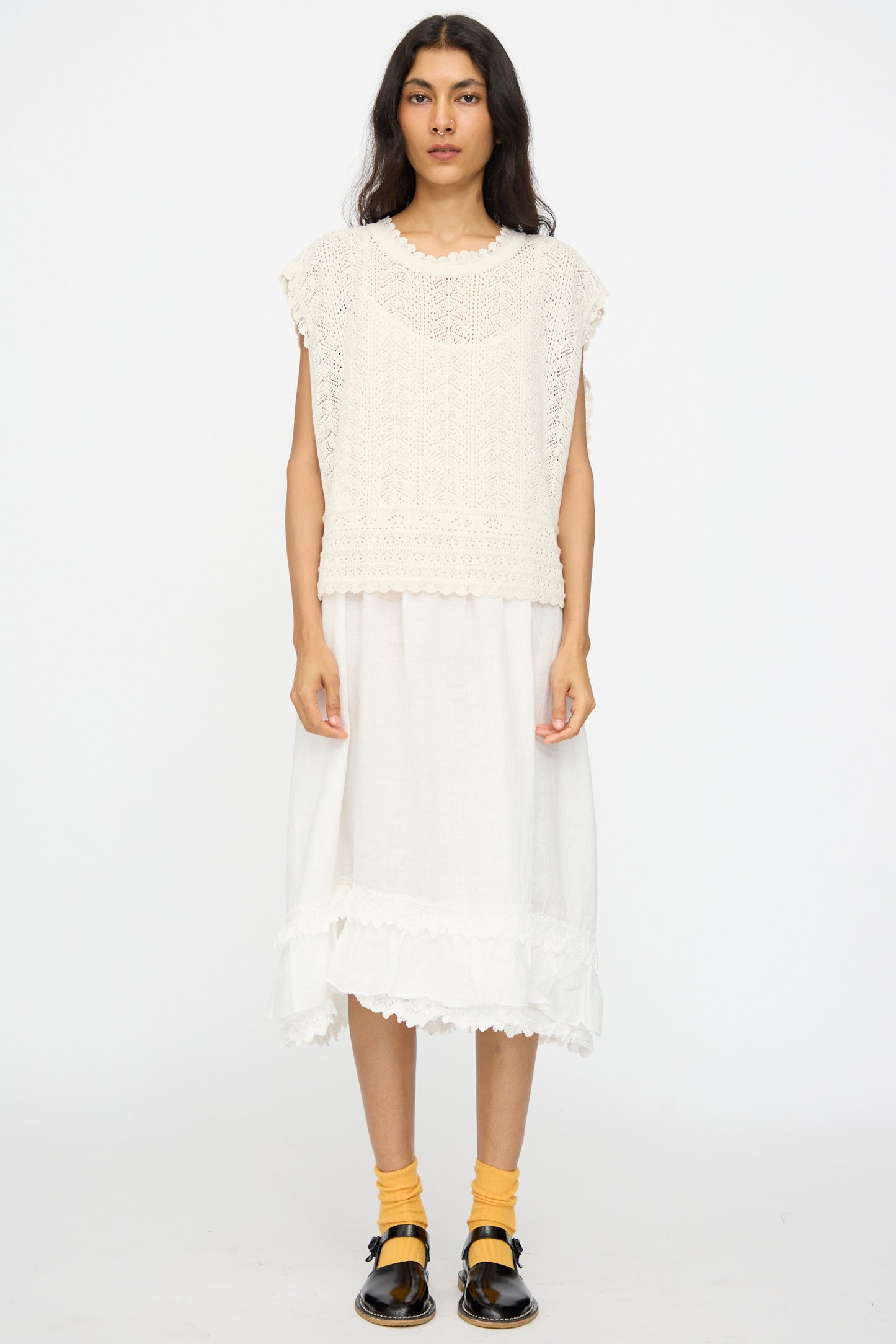 A person wears the nest Robe Openwork Lace Knit Vest in Off White over a white midi dress, paired with yellow socks and black Mary Jane shoes, against a white background.