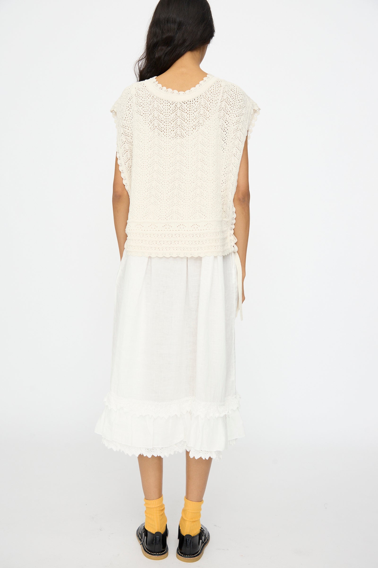 A woman faces away, wearing the nest Robe Openwork Lace Knit Vest in Off White over a white ruffle-trim skirt, paired with yellow socks and black sandals, set against a plain white background.