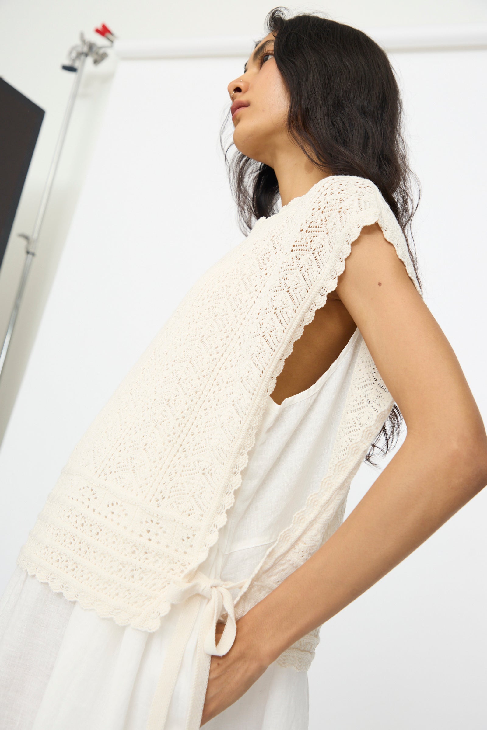 A woman wears the nest Robe Openwork Lace Knit Vest in Off White, a sleeveless cotton-linen blend with side ties, layered over a white dress against a white background.