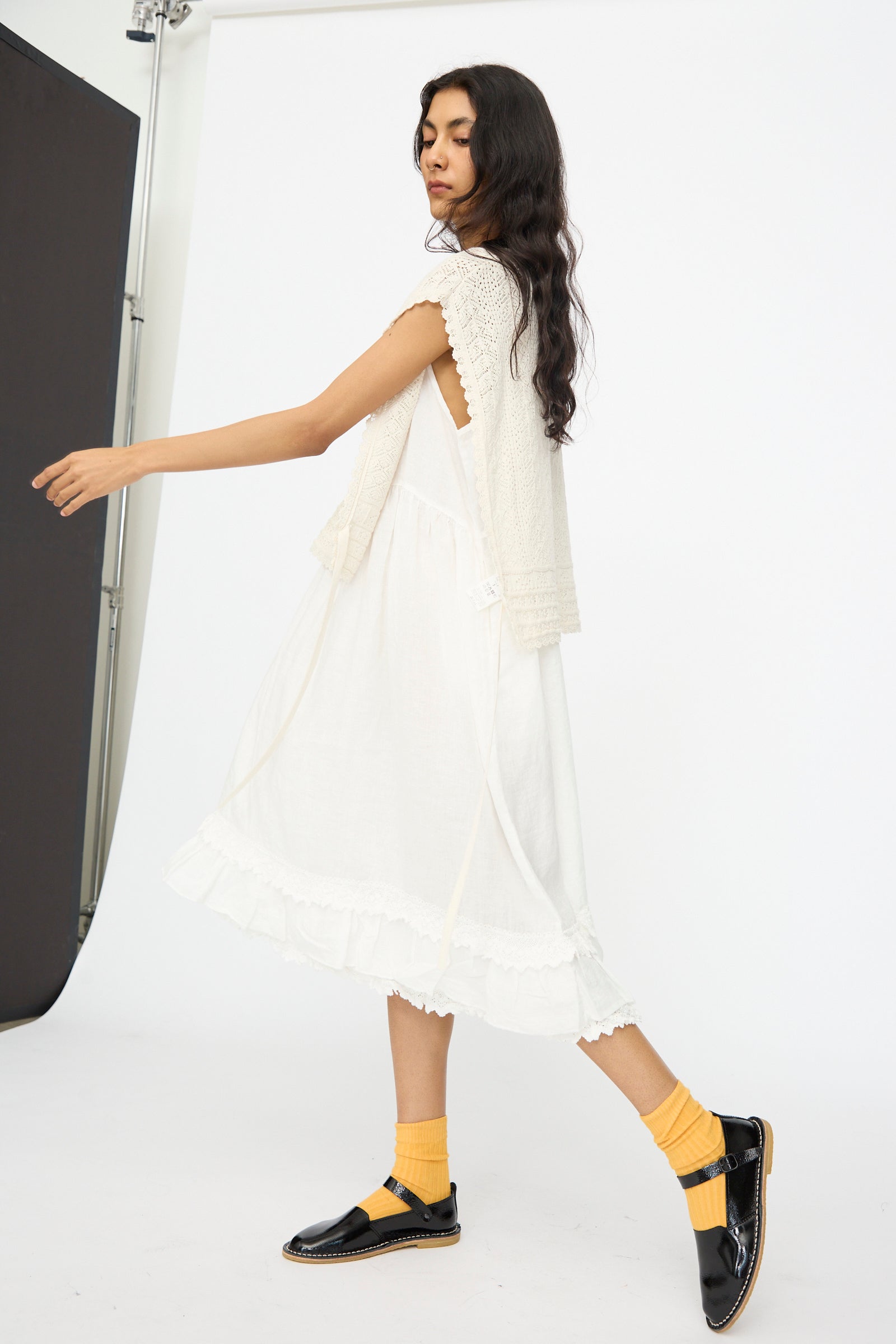A woman in a white dress, nest Robe’s Openwork Lace Knit Vest in Off White, yellow socks, and black shoes stands against a white studio background, looking over her shoulder.