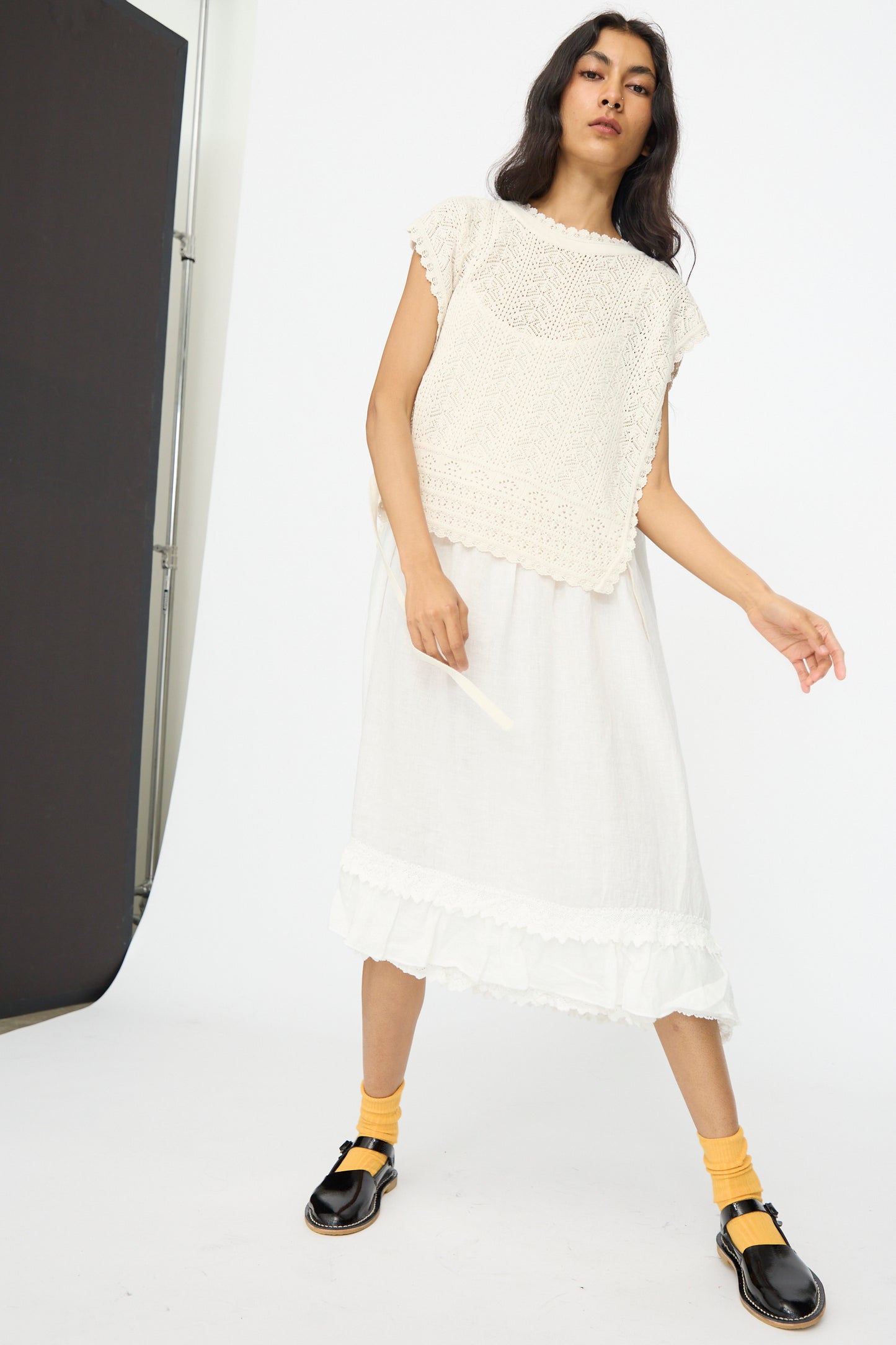 A woman stands in a studio wearing the nest Robe Openwork Lace Knit Vest in Off White, a white midi skirt, yellow socks, and black loafers, holding a small white item with a neutral expression.