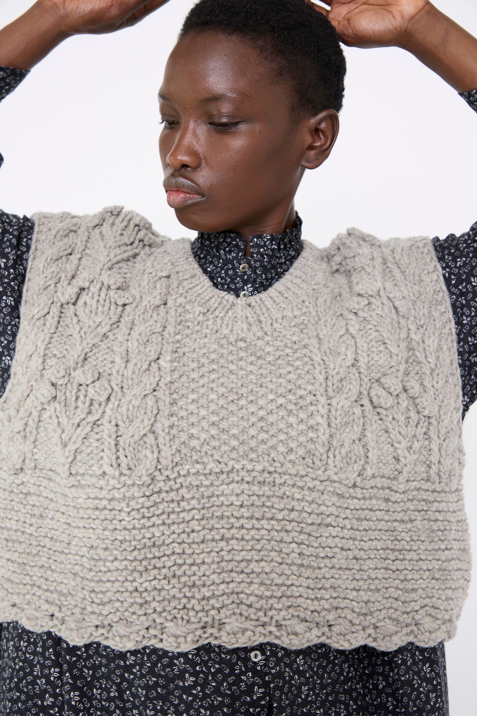 A person dressed in nest Robe's Peruvian Wool Bobble Knit Two-Way Vest in Beige over a patterned shirt, with arms raised and eyes looking to the side against a white background.