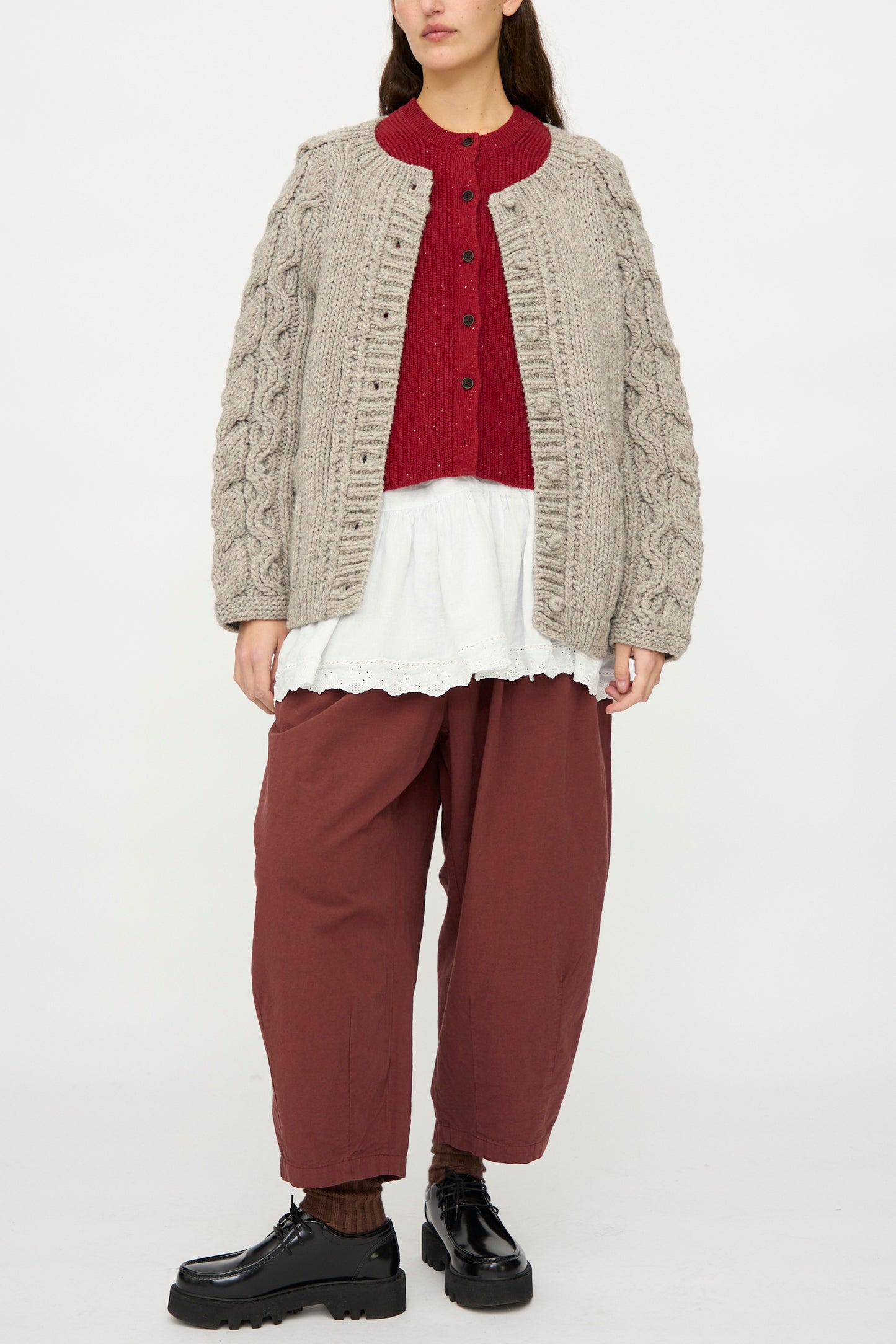 A person wears the nest Robe Peruvian Wool Cable Sleeve Knit Cardigan in Beige (oversized fit), a red button-up sweater, white tunic, loose maroon pants, brown socks, and black platform shoes, standing against a plain background.