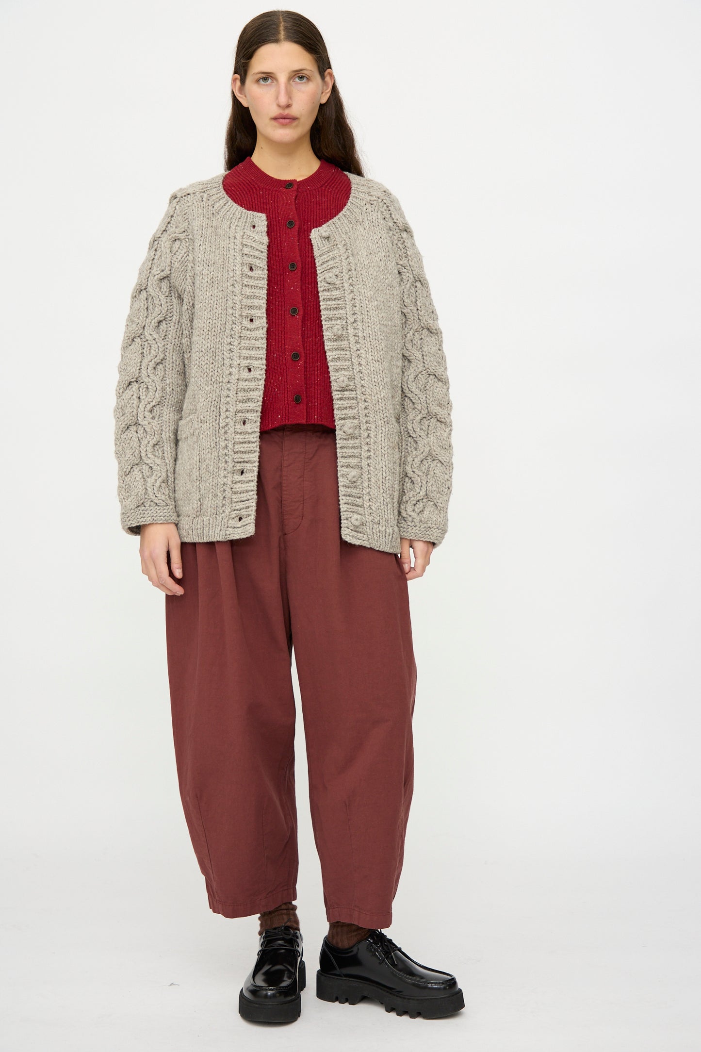 A person stands facing forward in a nest Robe Peruvian Wool Cable Sleeve Knit Cardigan in Beige, paired with a red buttoned sweater, loose maroon pants, and black platform shoes against a plain white background.