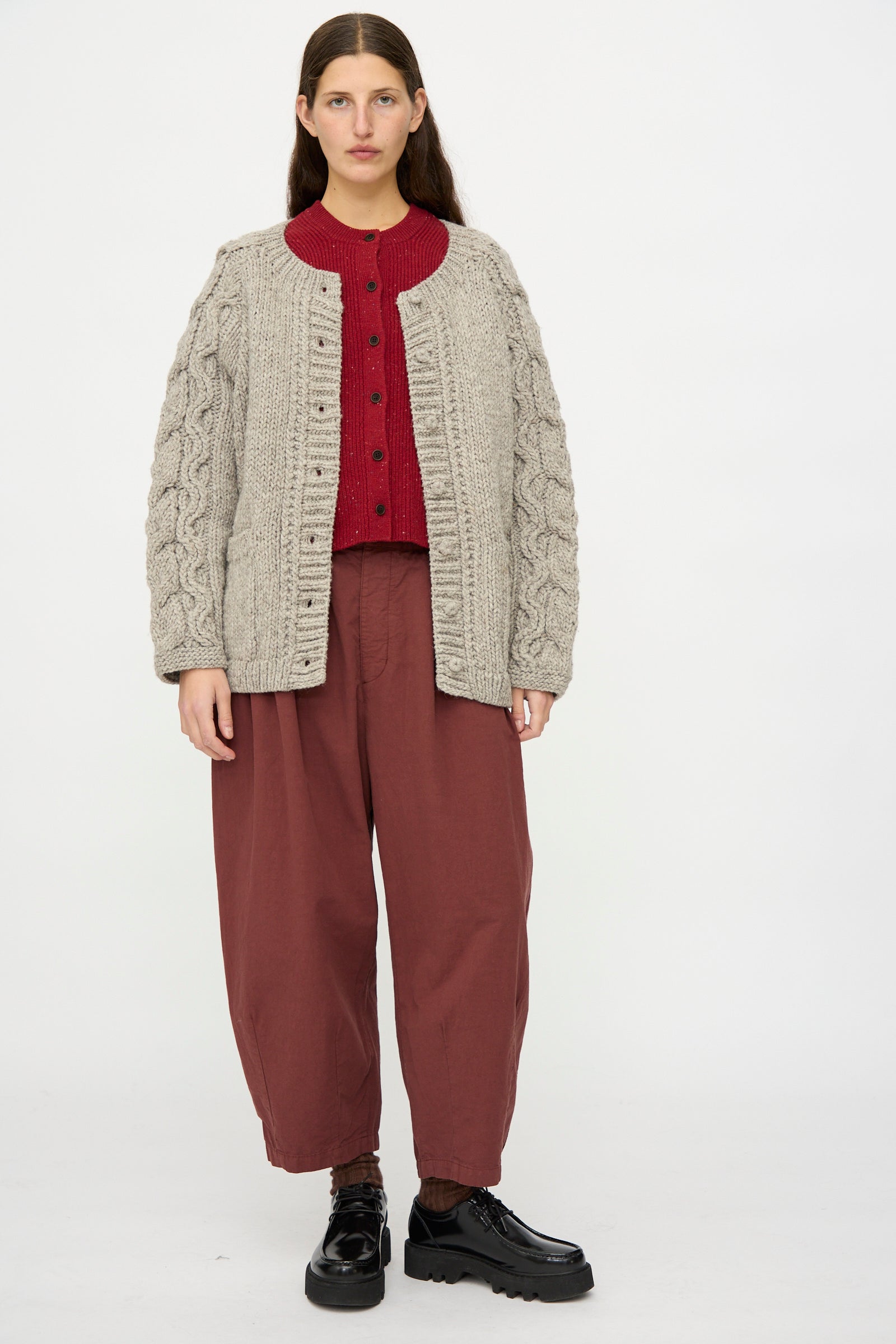 A person stands facing forward in a nest Robe Peruvian Wool Cable Sleeve Knit Cardigan in Beige, paired with a red buttoned sweater, loose maroon pants, and black platform shoes against a plain white background.