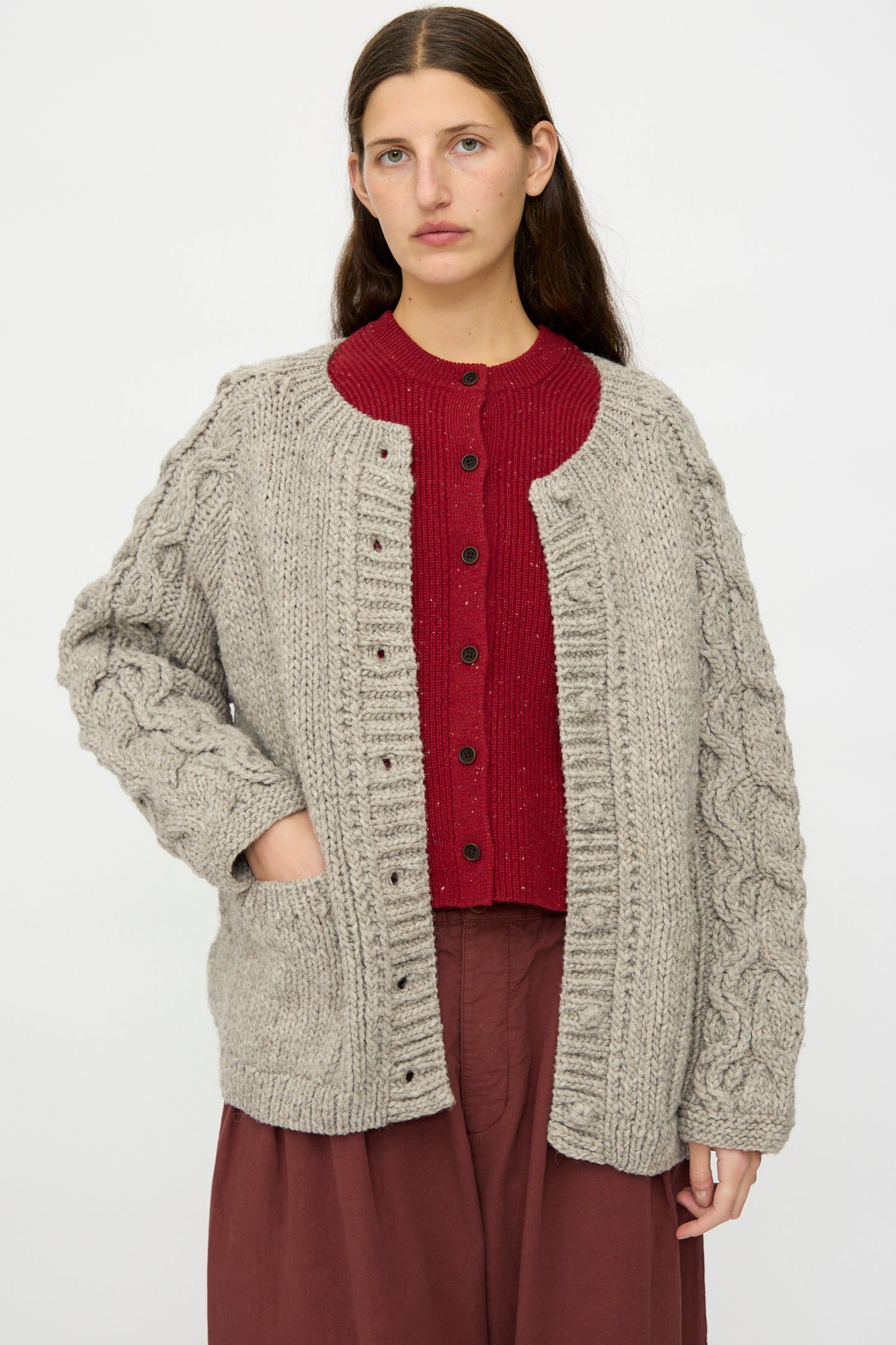 A woman with long brown hair wears the nest Robe Peruvian Wool Cable Sleeve Knit Cardigan in Beige over a red buttoned sweater and maroon skirt, standing against a plain white background.