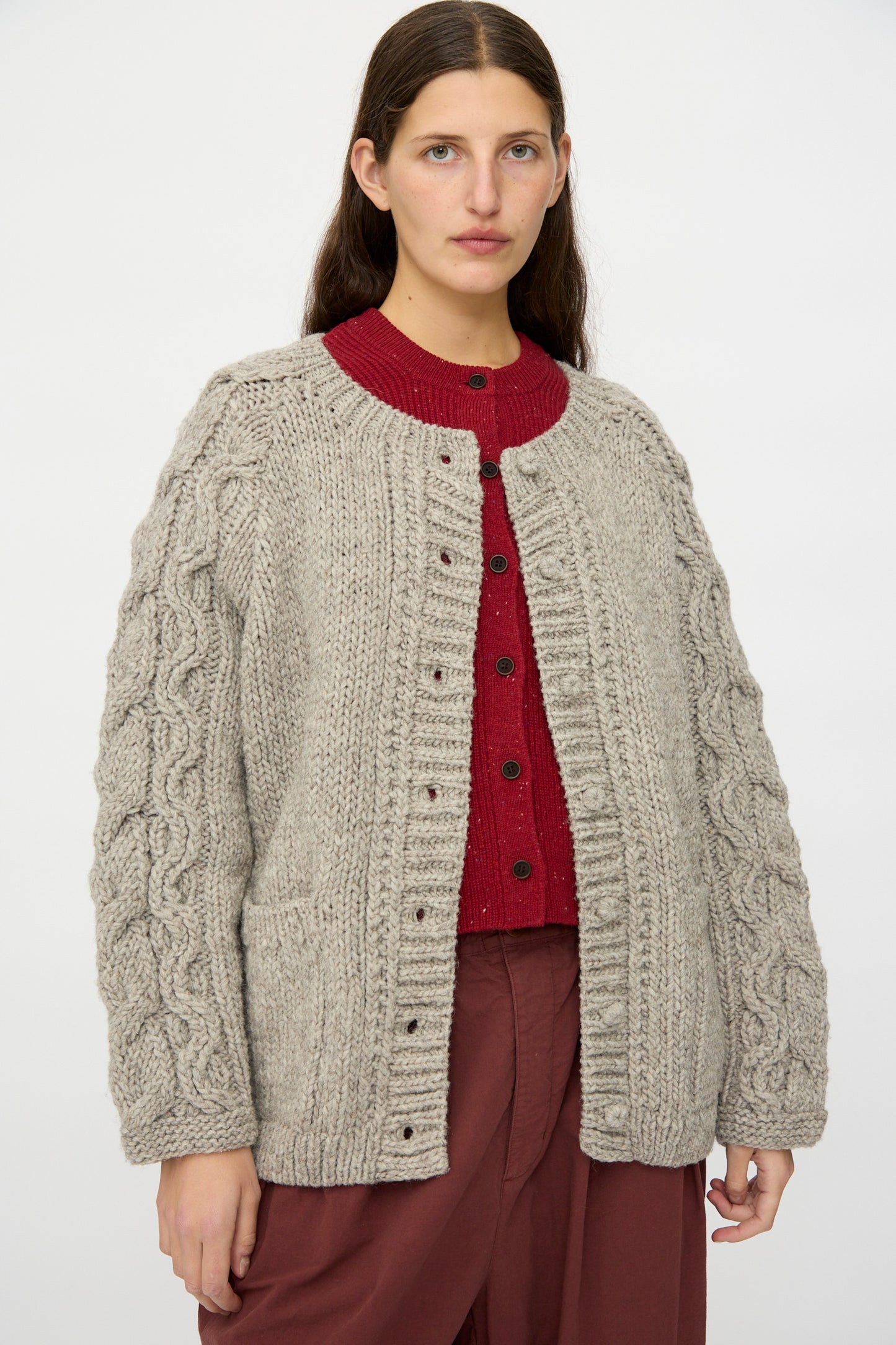 A person wearing a nest Robe Peruvian Wool Cable Sleeve Knit Cardigan in Beige over a red sweater and maroon pants, standing against a plain white background.